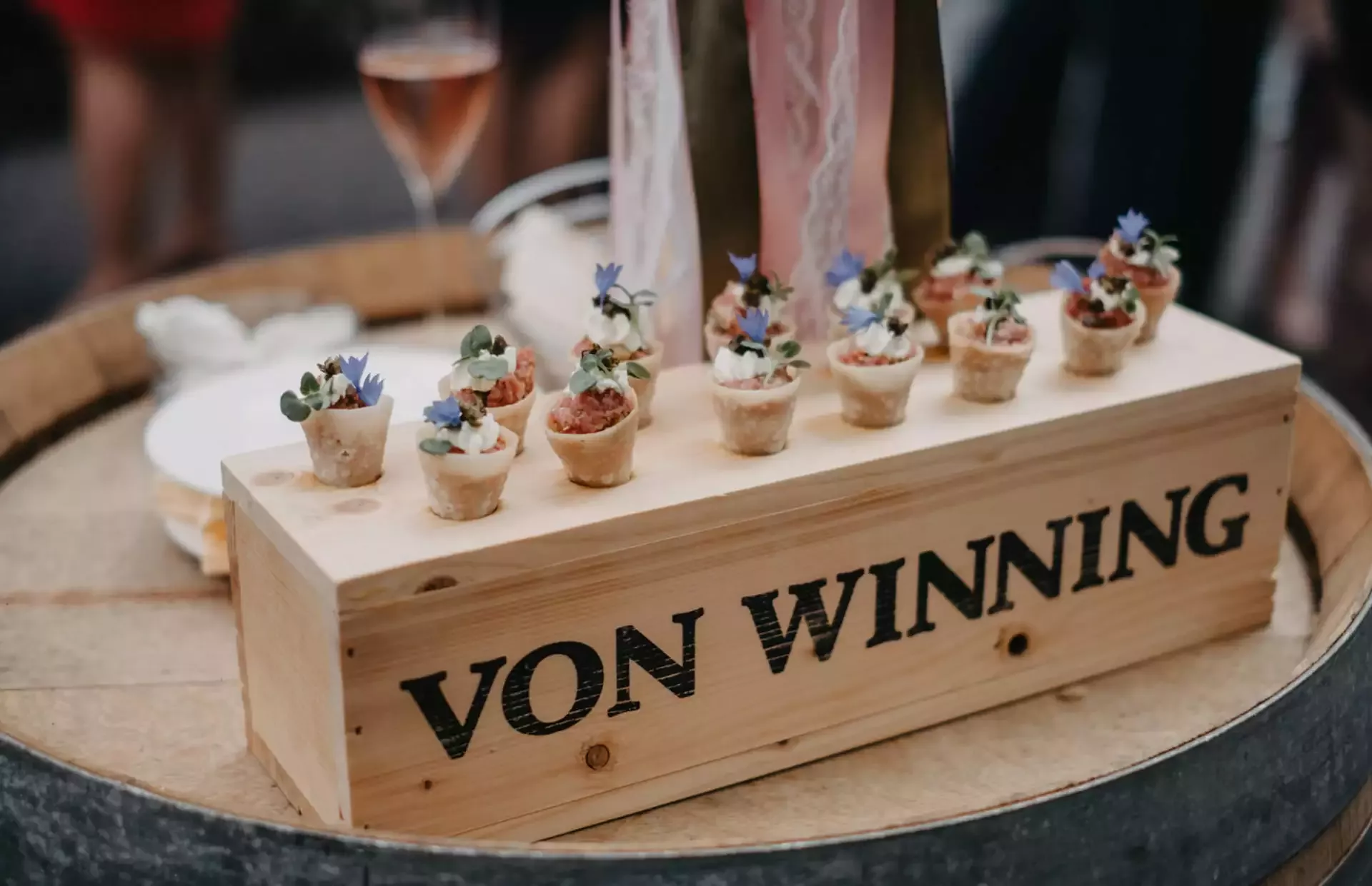 Auf einer Holzkiste mit der Aufschrift „Von Winning“ auf einem Fass werden Mini-Häppchen mit Garnitur präsentiert. Im Hintergrund steht ein Glas mit rosafarbenem Getränk, dahinter sind Teller und Menschen leicht zu erkennen.