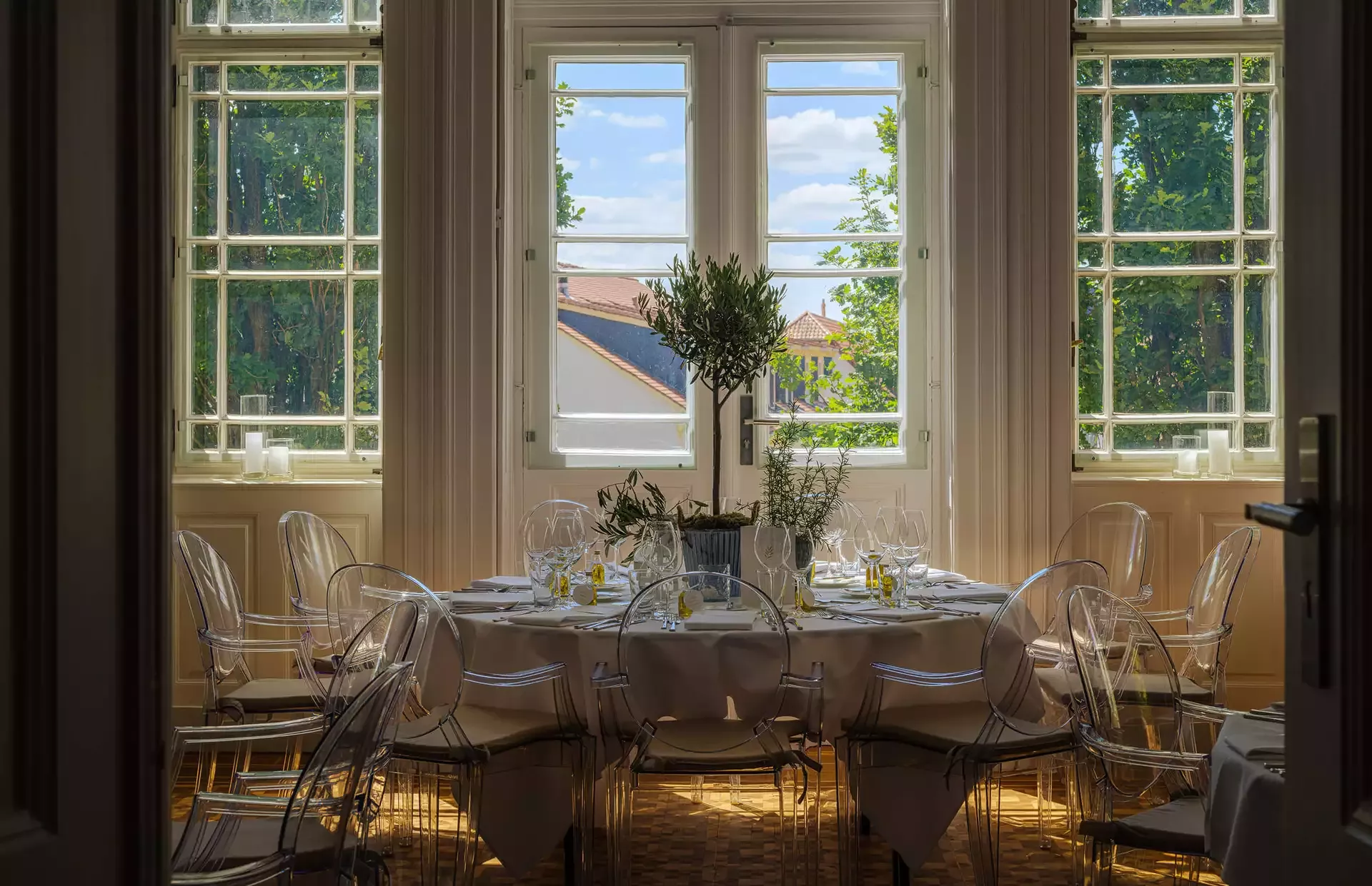 Elegantes Esszimmer mit einem runden, für eine Mahlzeit gedeckten Tisch mit transparenten Stühlen und Topfpflanzen als Tafelaufsatz. Große Fenster bieten einen Blick auf grüne Bäume und einen klaren blauen Himmel und füllen den Raum mit natürlichem Licht.