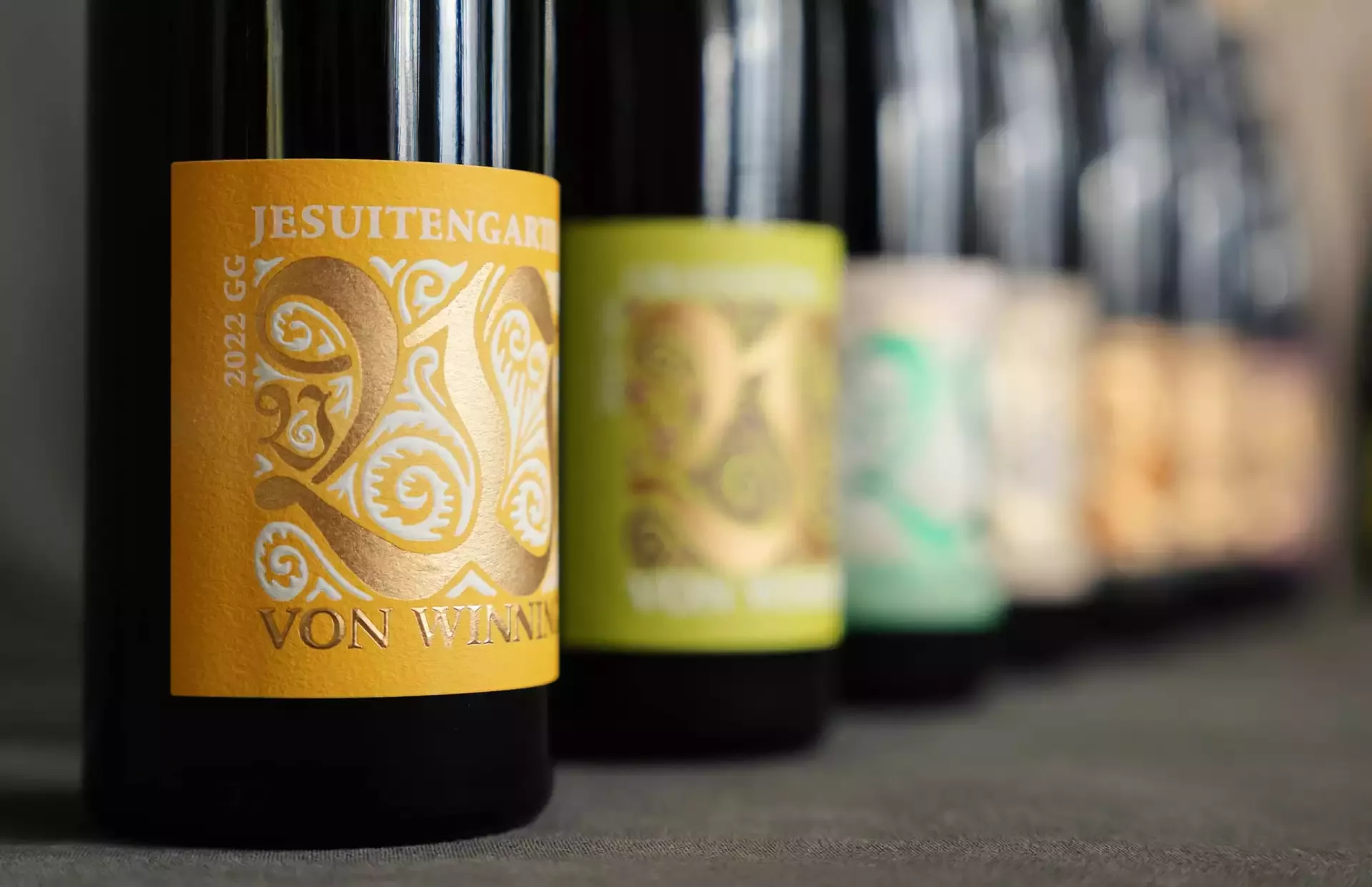 Nahaufnahme einer Reihe von Weinflaschen mit bunten Etiketten. Die nächste Flasche hat ein gelbes Etikett mit dem Text „Jesuitengarten“ und „Von Winning“ in dekorativem Aufdruck, während die anderen Flaschen im Hintergrund verschwimmen.