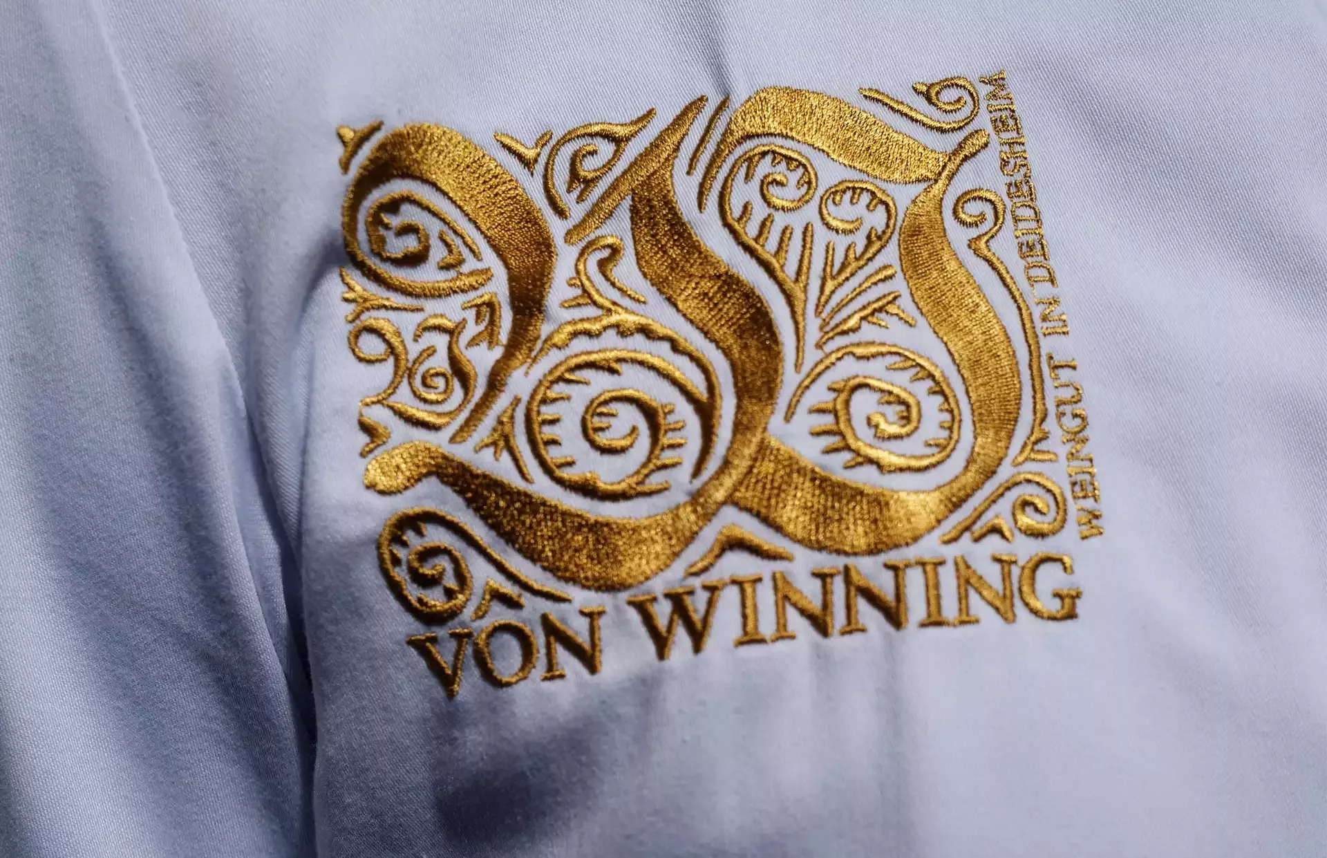 Eine Nahaufnahme eines goldgestickten Monogramms mit kunstvollen Mustern auf weißem Stoff. Darunter steht in Gold „VON WINNING“. Die Worte „WEINGUT IN DEIDESHEIM“ sind an der Seite sichtbar.