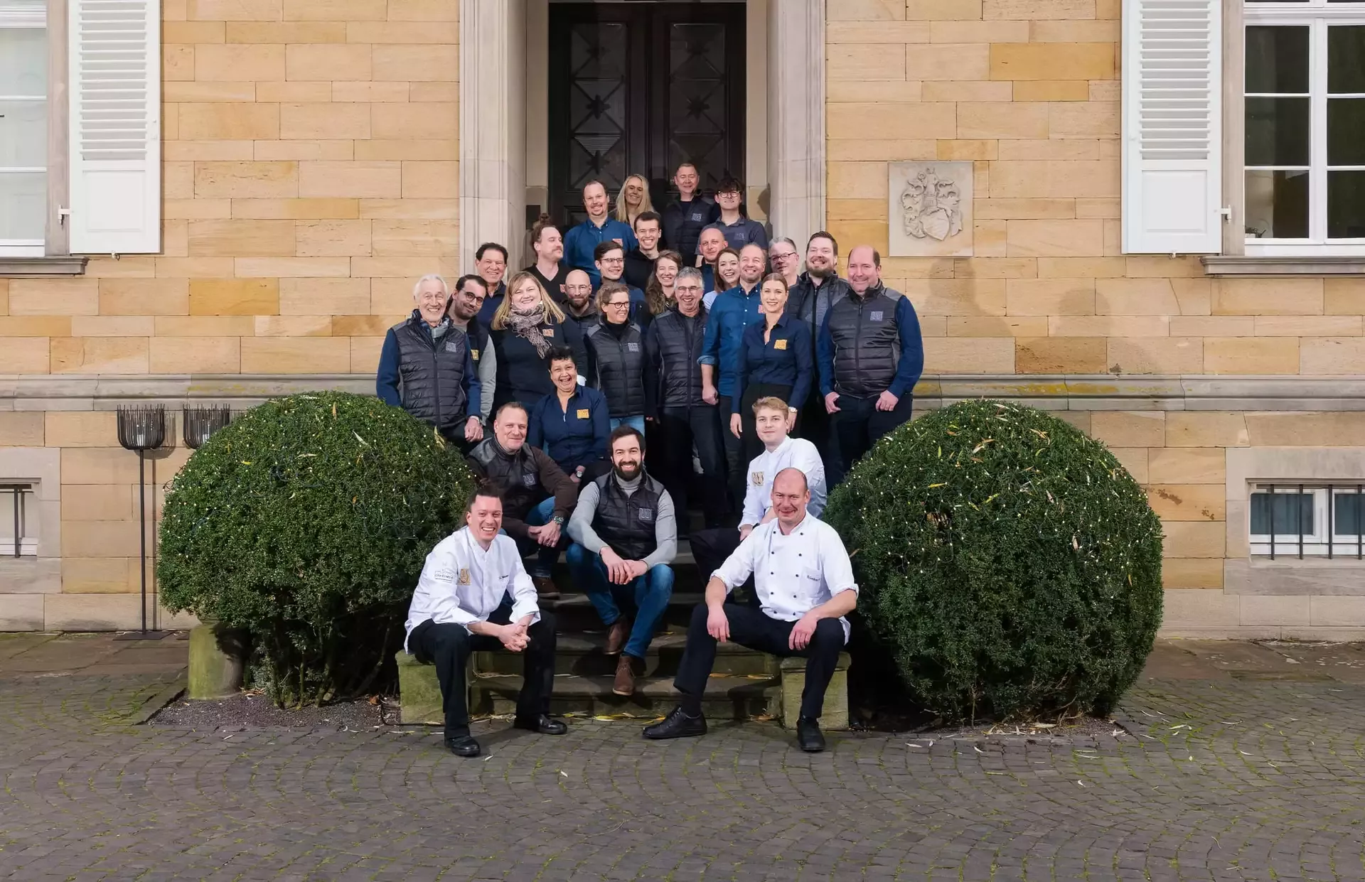 Eine Gruppe von Menschen posiert auf Steinstufen vor einem großen Gebäude mit Holztüren. Die meisten tragen dunkle Jacken, und drei Personen in der ersten Reihe tragen Kochuniformen. Zwei gestutzte Büsche flankieren die Gruppe.