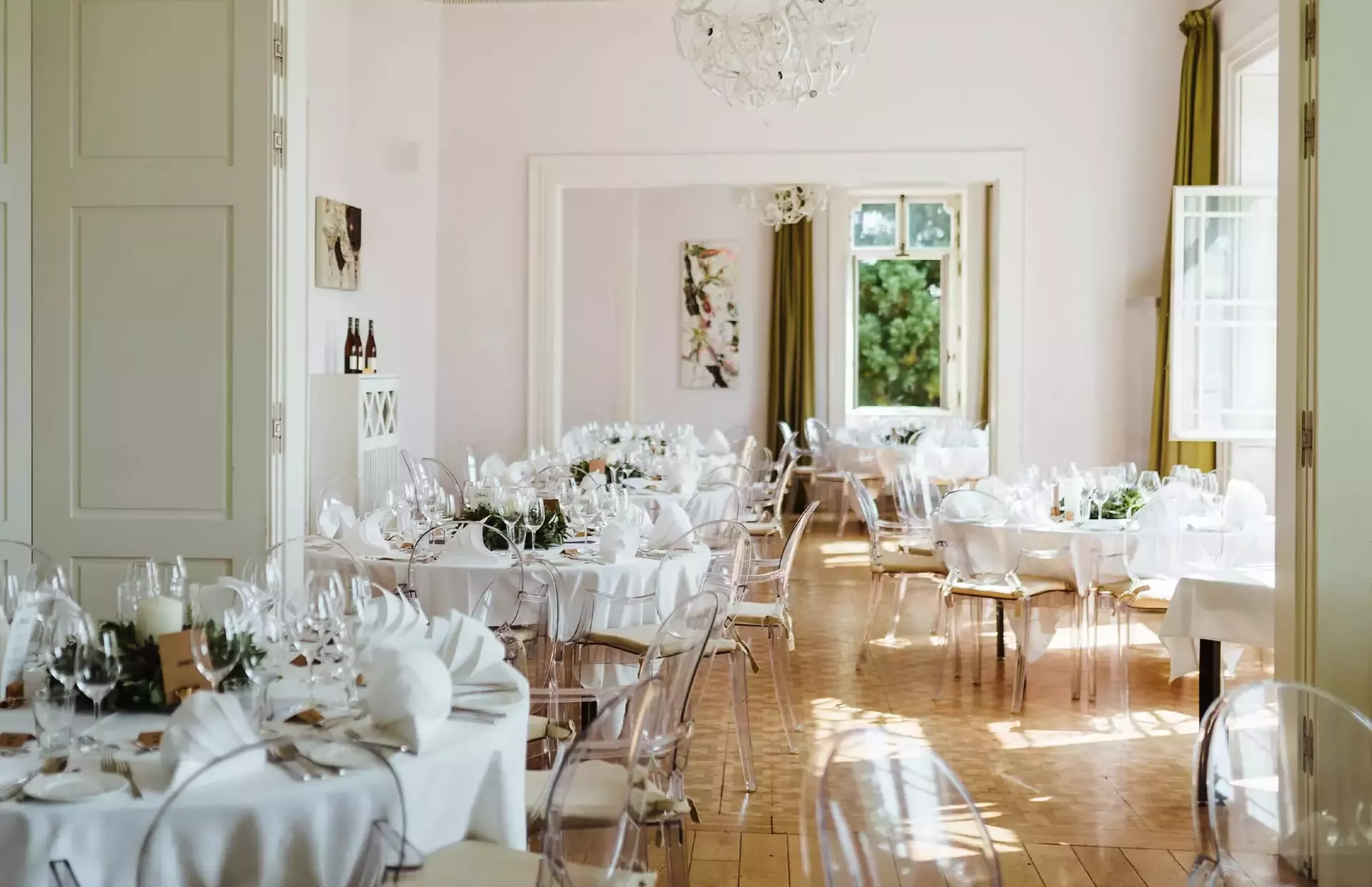 Eleganter, sonnendurchfluteter Speisesaal für eine Veranstaltung mit runden, mit weißen Tischdecken bedeckten Tischen, transparenten Stühlen und ordentlich arrangiertem Gedeck. Zur Dekoration gehören Blumengestecke und Weinflaschen. Große Fenster lassen natürliches Licht herein.