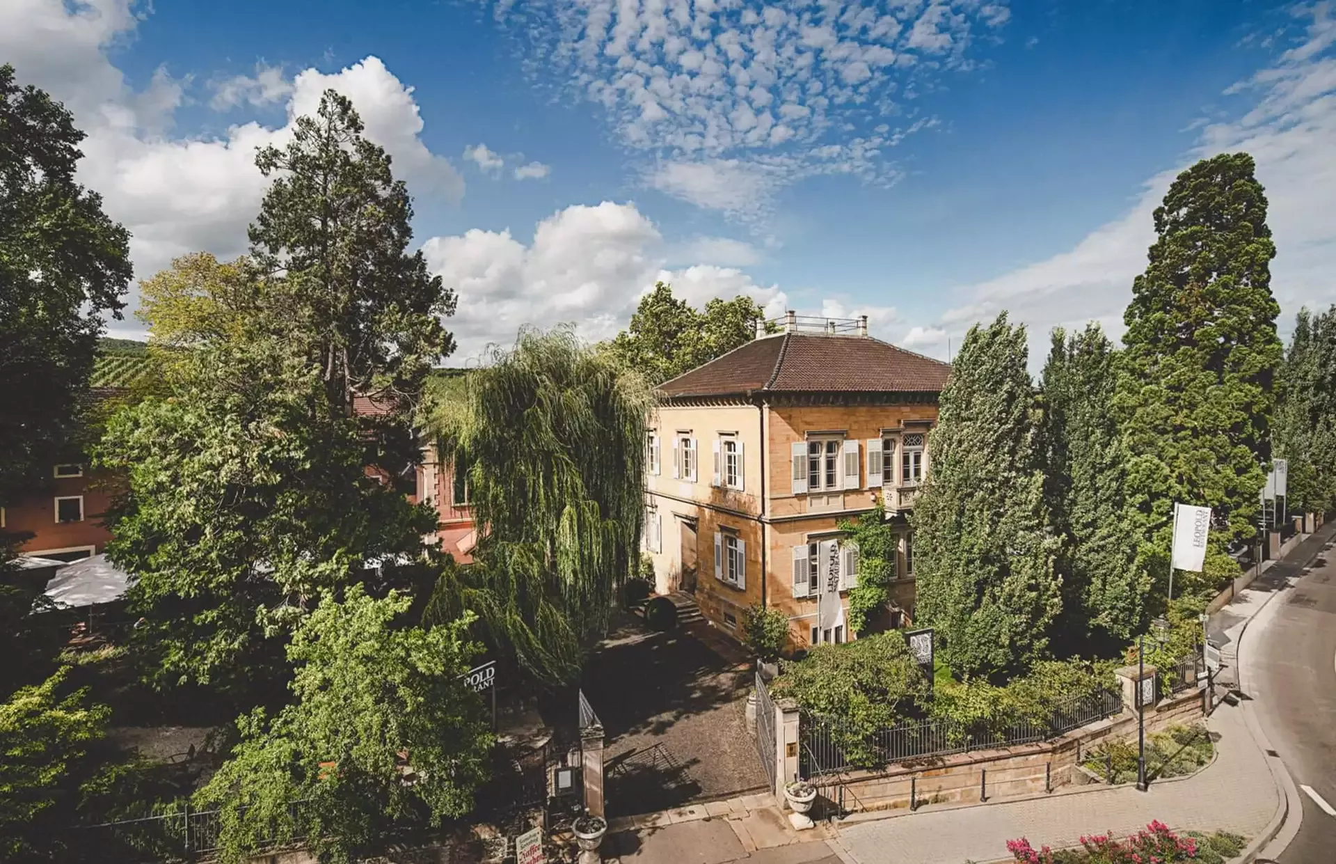 Ein malerischer Blick auf ein historisches gelbes Backsteingebäude, umgeben von üppigem Grün und Bäumen. Ein gepflasterter Weg führt zum Eingang und der Himmel darüber ist mit flauschigen Wolken übersät. Eine Straße führt rechts um das Grundstück herum.