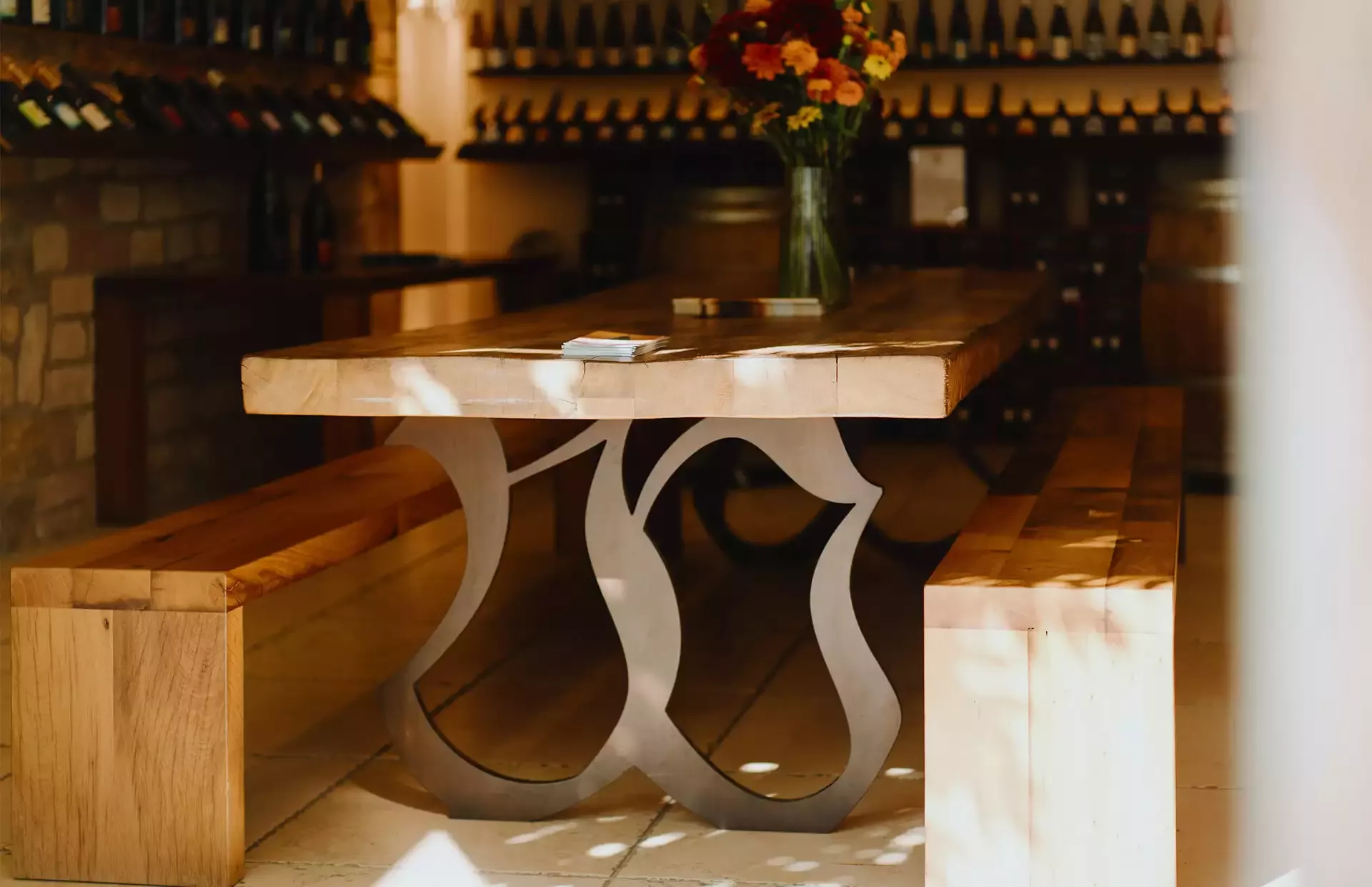 Ein Holztisch mit einem kunstvollen Metallgestell steht in einem Weinkeller. Zwei Holzbänke flankieren den Tisch. Darauf steht eine Vase mit bunten Blumen. Sonnenlicht und Schatten erzeugen Muster auf dem Boden und den Möbeln. Im Hintergrund stehen Weinflaschen.