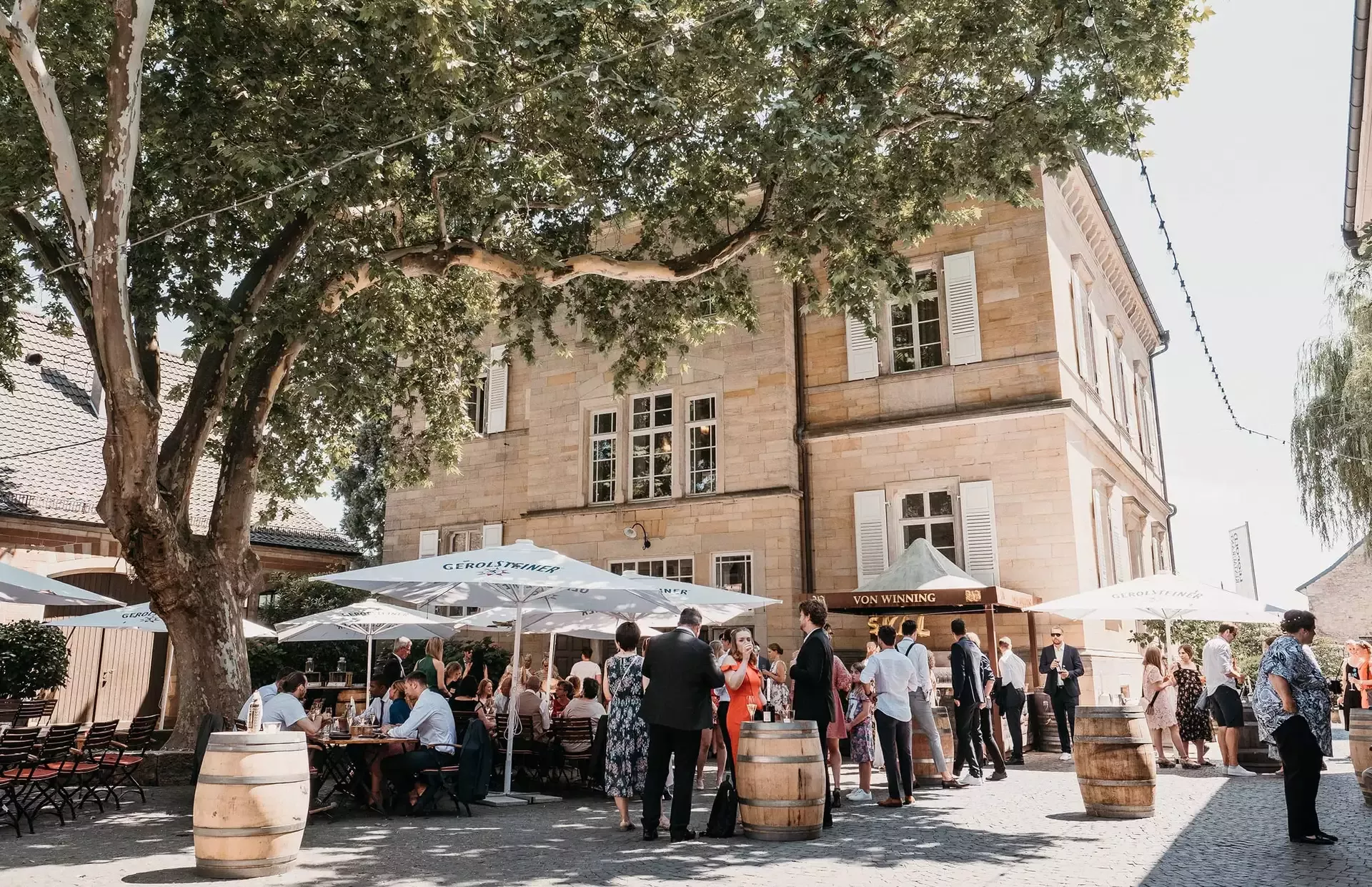 Menschen treffen sich im Freien in einem Restaurant mit schattigen Tischen unter Sonnenschirmen. Die Kulisse besteht aus einem großen Baum und einem historischen Steingebäude. Zur Dekoration gehören Lichterketten und in der Nähe stehen Holzfässer. Die Szene lässt auf eine gesellige oder festliche Veranstaltung schließen.