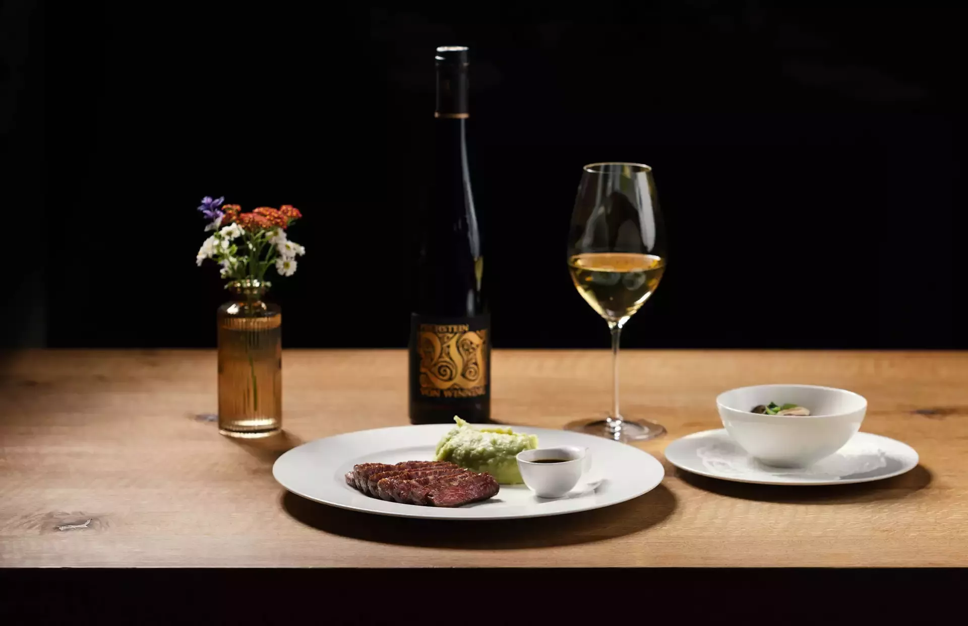 Ein Esstisch auf einem Holztisch besteht aus einem Teller mit Steak, Soße und Gemüse, einer Schüssel Suppe, einer Flasche Wein und einem mit Weißwein gefüllten Weinglas. Neben der Weinflasche steht eine kleine Vase mit bunten Blumen.