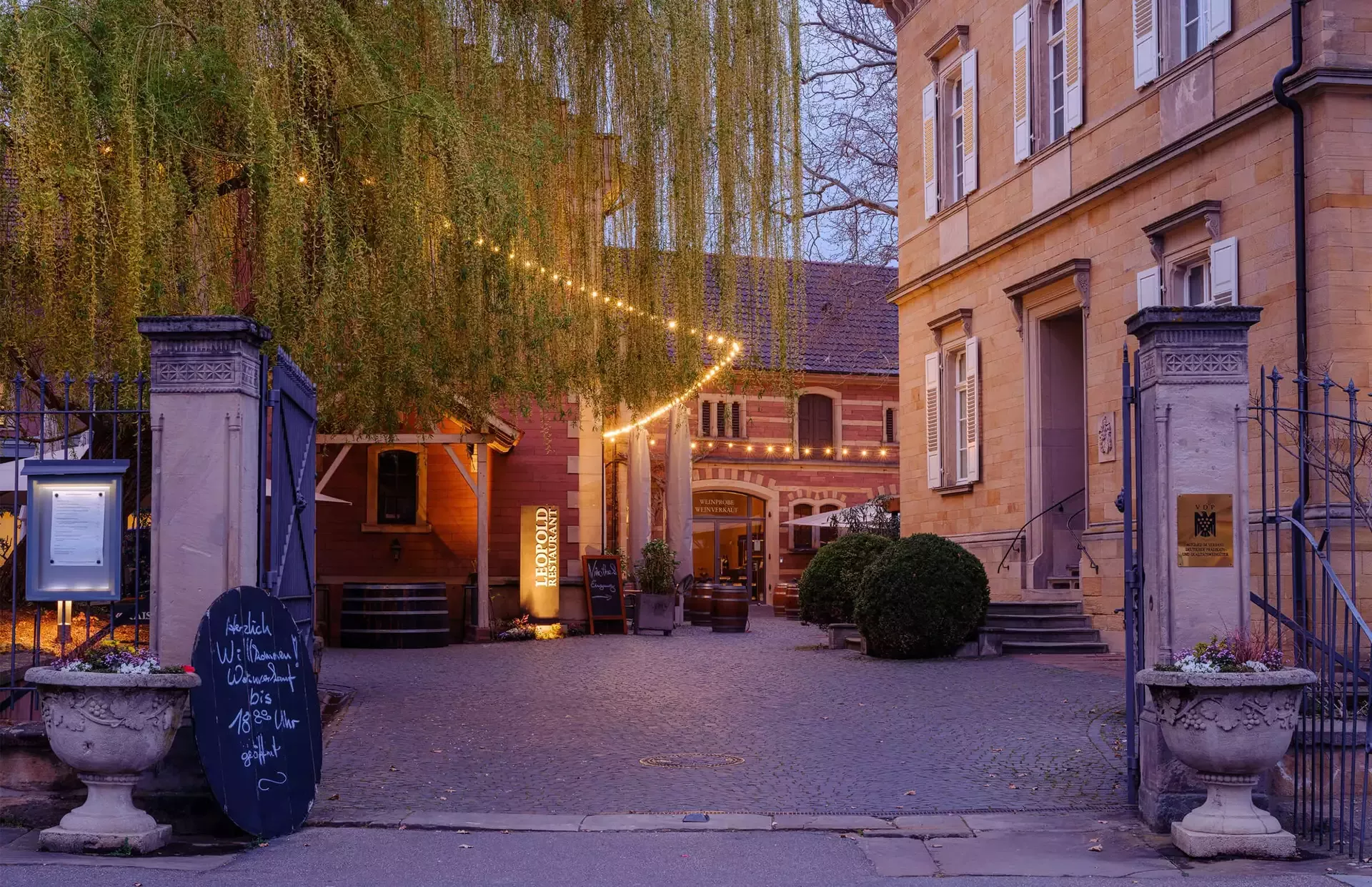 Ein charmanter Innenhof mit Kopfsteinpflaster und Gebäuden mit Bogenfenstern. Ein Baum mit Lichterketten und eine Tafel mit Text verleihen Charakter. Warmes Licht schafft eine gemütliche Atmosphäre und Topfpflanzen schmücken den Eingang.