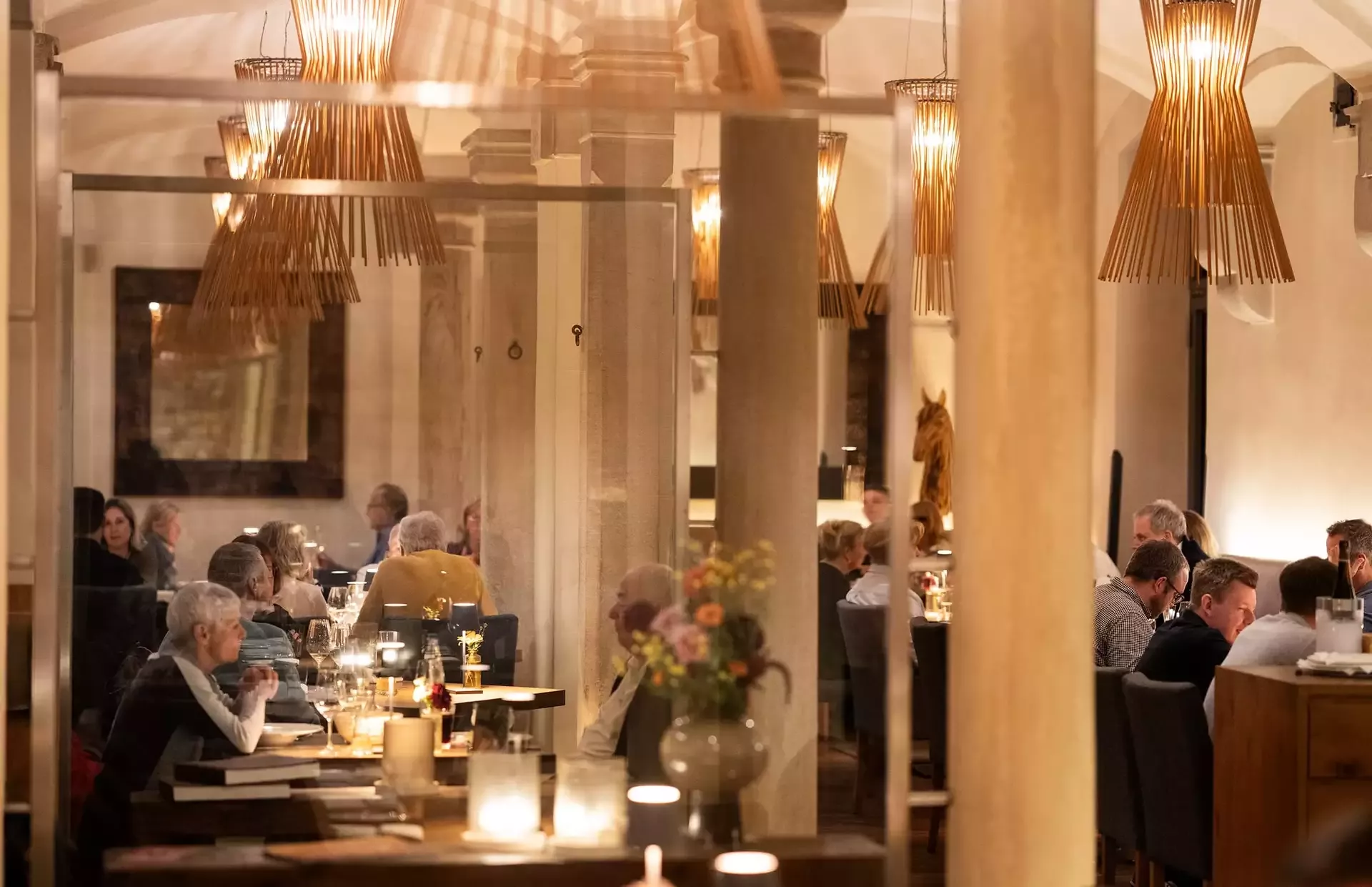 Ein warm beleuchtetes Restaurantinneres mit Leuten, die an Tischen speisen. Der Raum ist mit hohen Holzlampen und Blumenarrangements ausgestattet. Die Gäste unterhalten sich, was eine gemütliche und einladende Atmosphäre schafft.