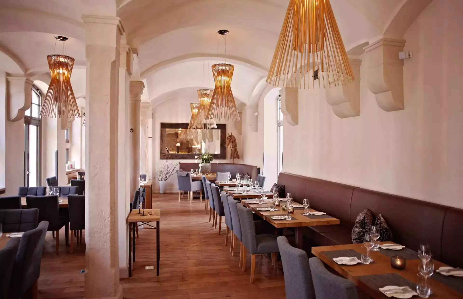 Ein modernes, elegantes Restaurant-Interieur mit gewölbten Decken und Holzböden. Mehrere Tische mit blauen Stühlen und Gläsern und Servietten. Warmes Licht von stilvollen Kronleuchtern sorgt für eine gemütliche Atmosphäre.