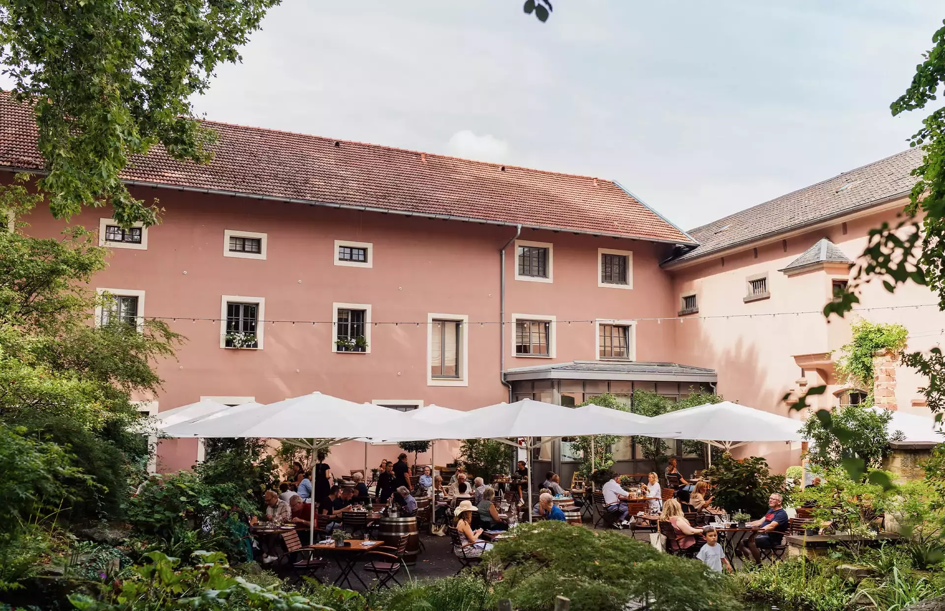 Menschen genießen Essen und Getränke auf der Außenterrasse des Restaurant Leopold, die von großen weißen Sonnenschirmen beschattet wird. Die Umgebung ist von Grün umgeben und grenzt an ein rustikales, pfirsichfarbenes Gebäude mit mehreren Fenstern. Die Atmosphäre ist lebhaft und entspannt.