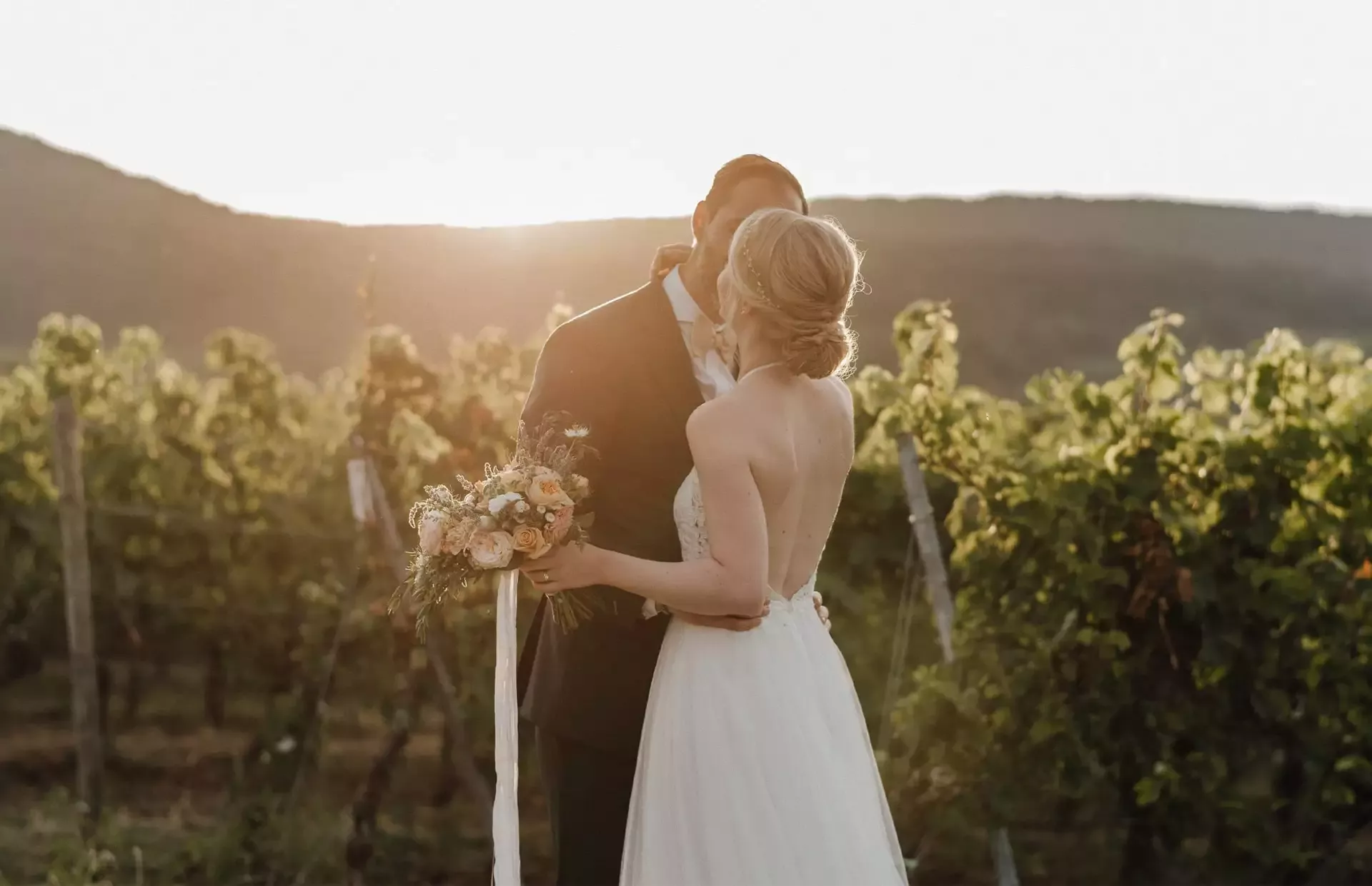 Braut und Bräutigam küssen sich bei Sonnenuntergang in einem Weinberg. Die Braut trägt ein schulterfreies Kleid und hält einen Blumenstrauß in der Hand. Der Bräutigam trägt einen dunklen Anzug. Die Sonne wirft einen warmen Schein auf die üppigen grünen Weinreben im Hintergrund.