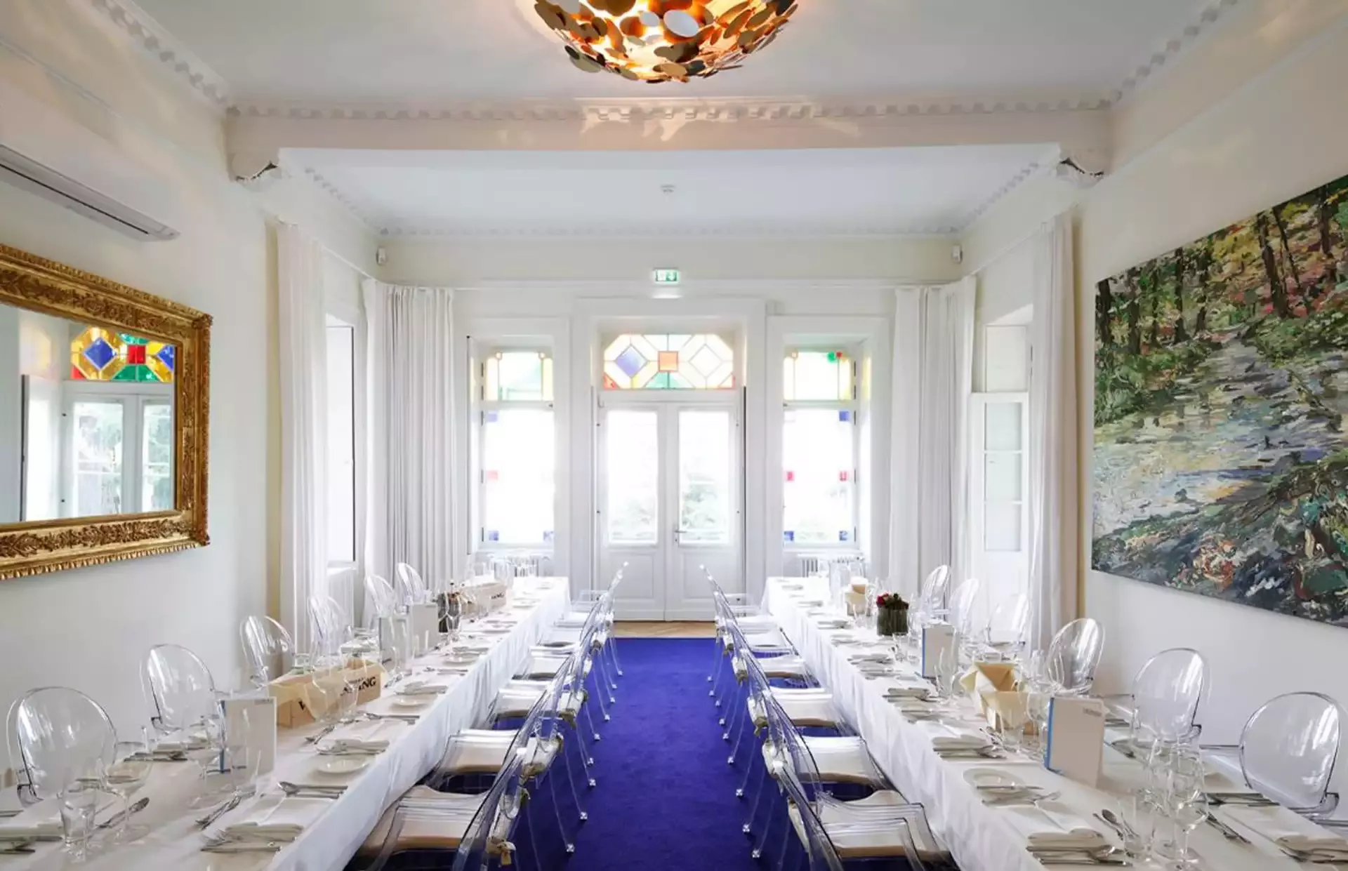 Ein langer, eleganter Speisesaal mit weißen Wänden und einem violetten Teppich. Er verfügt über einen großen Tisch mit weißen Tischdecken, durchsichtigen Stühlen und Geschirr. Bunte Buntglasfenster und eine kunstvolle Deckenleuchte werten das Ambiente auf.