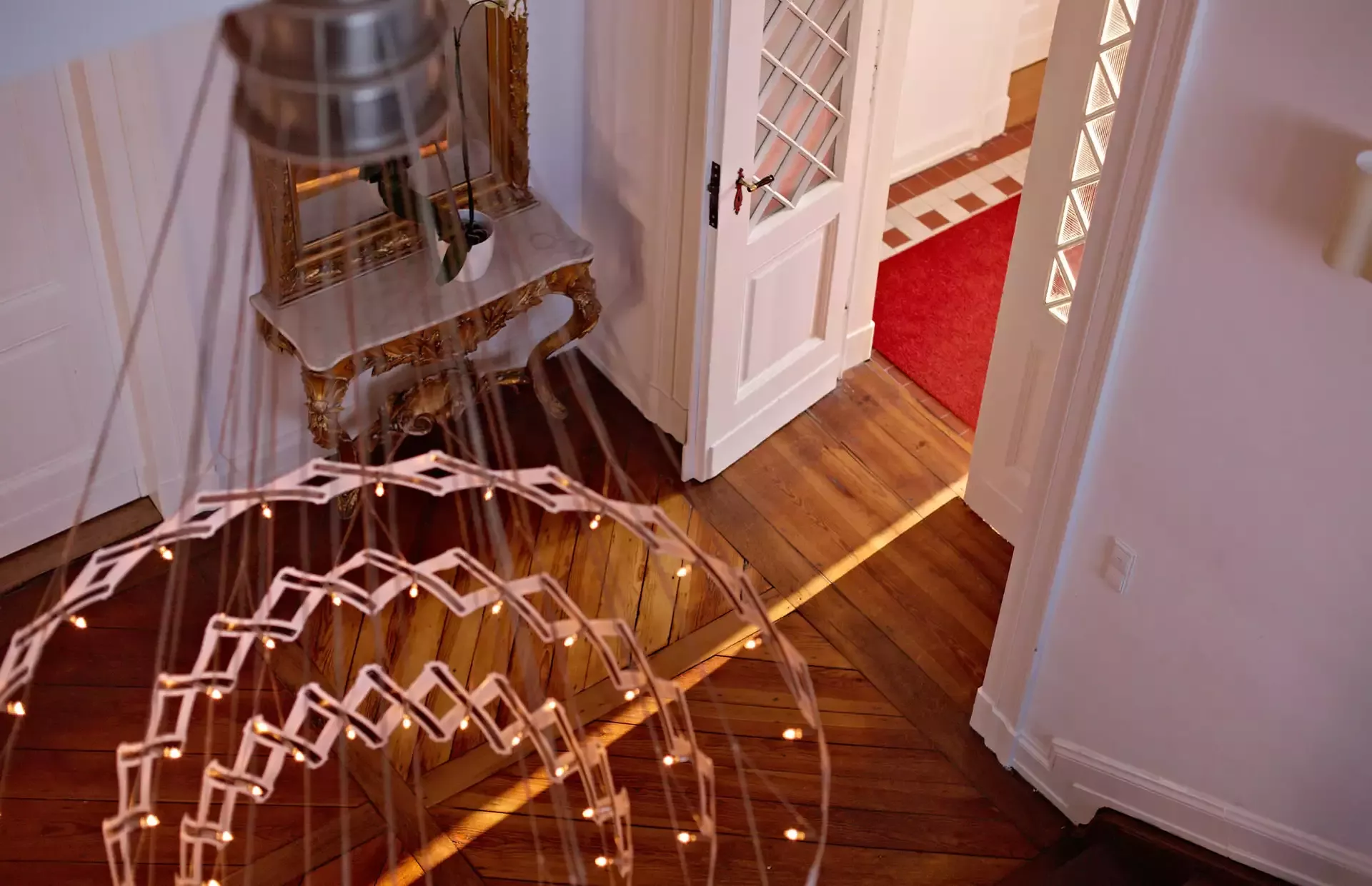Blick von oben auf ein stilvolles Interieur mit Holzböden. Im Vordergrund hängt ein geometrischer Kronleuchter. Eine offene Tür gibt den Blick auf einen Raum mit rotem Teppich und einem dekorativen goldenen Konsolentisch frei. Die Wände sind weiß mit eleganten Details.