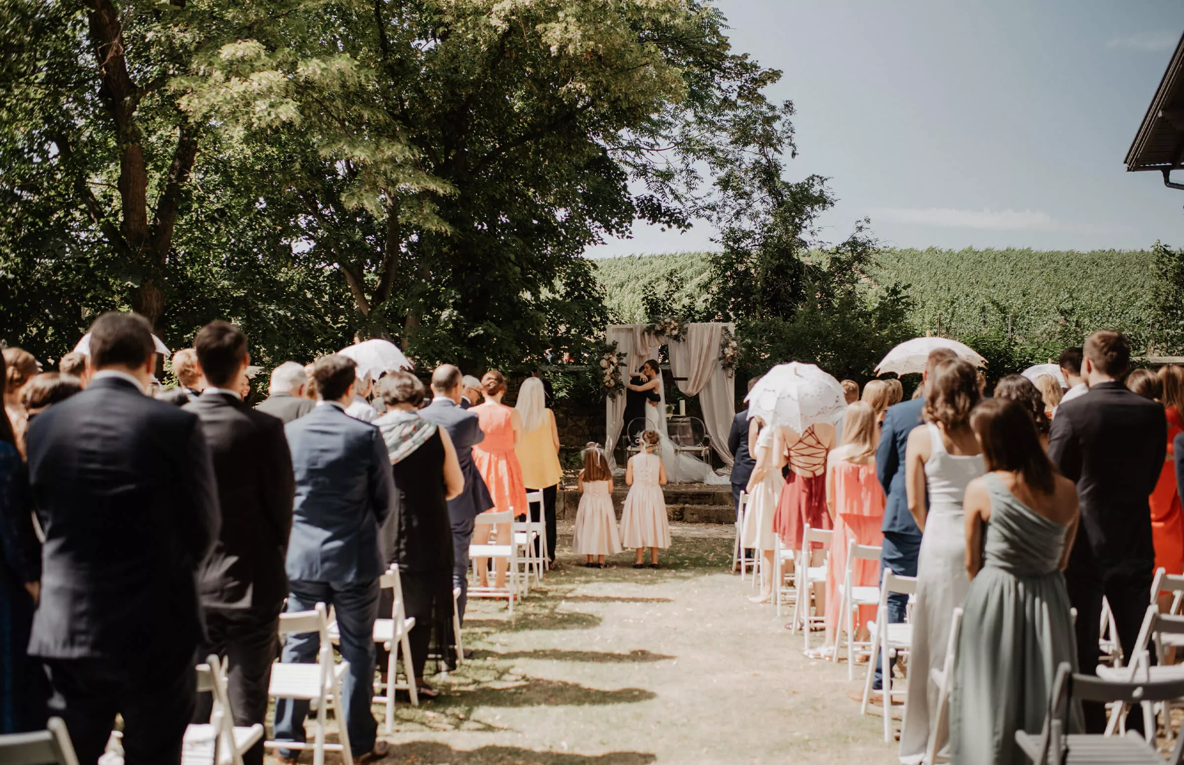 Eine Hochzeitszeremonie in einem Garten im Freien. Gäste in farbenfroher Kleidung sitzen entlang eines mit Gras bewachsenen Ganges. Das Paar steht unter einem Baldachin am Altar, umgeben von Grün und Bäumen. Im Hintergrund sind ein sonniger Himmel und ein offenes Feld zu sehen.