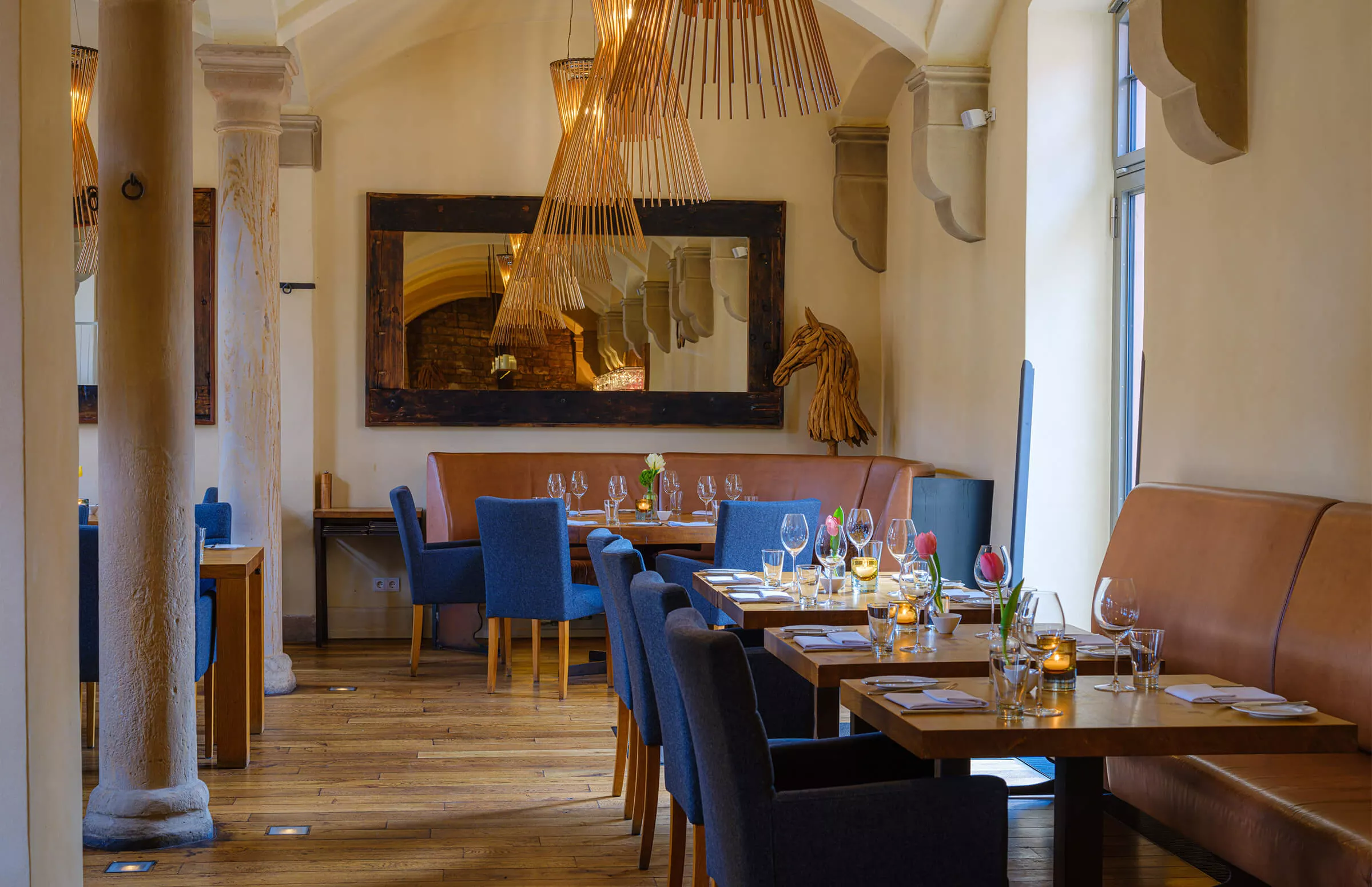 Ein gemütliches Restaurant-Interieur mit Holzfußboden, blau gepolsterten Stühlen und braunen Lederbänken. Die Tische sind mit Weingläsern, Kerzen und Blumen gedeckt. Warmes Licht und ein großer Spiegel verstärken die einladende Atmosphäre.