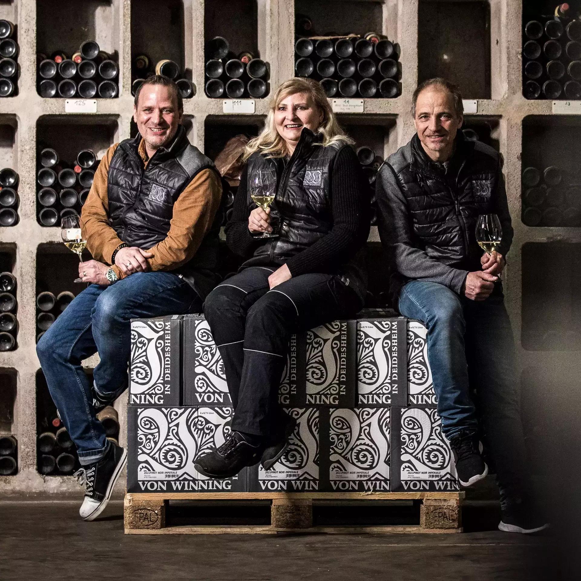 Drei Personen in passenden Westen sitzen auf Weinkisten in einem Keller, umgeben von Regalen voller Weinflaschen. Jeder von ihnen hält ein Glas Wein in der Hand und lächelt in die Kamera, wodurch eine warme und einladende Atmosphäre entsteht.