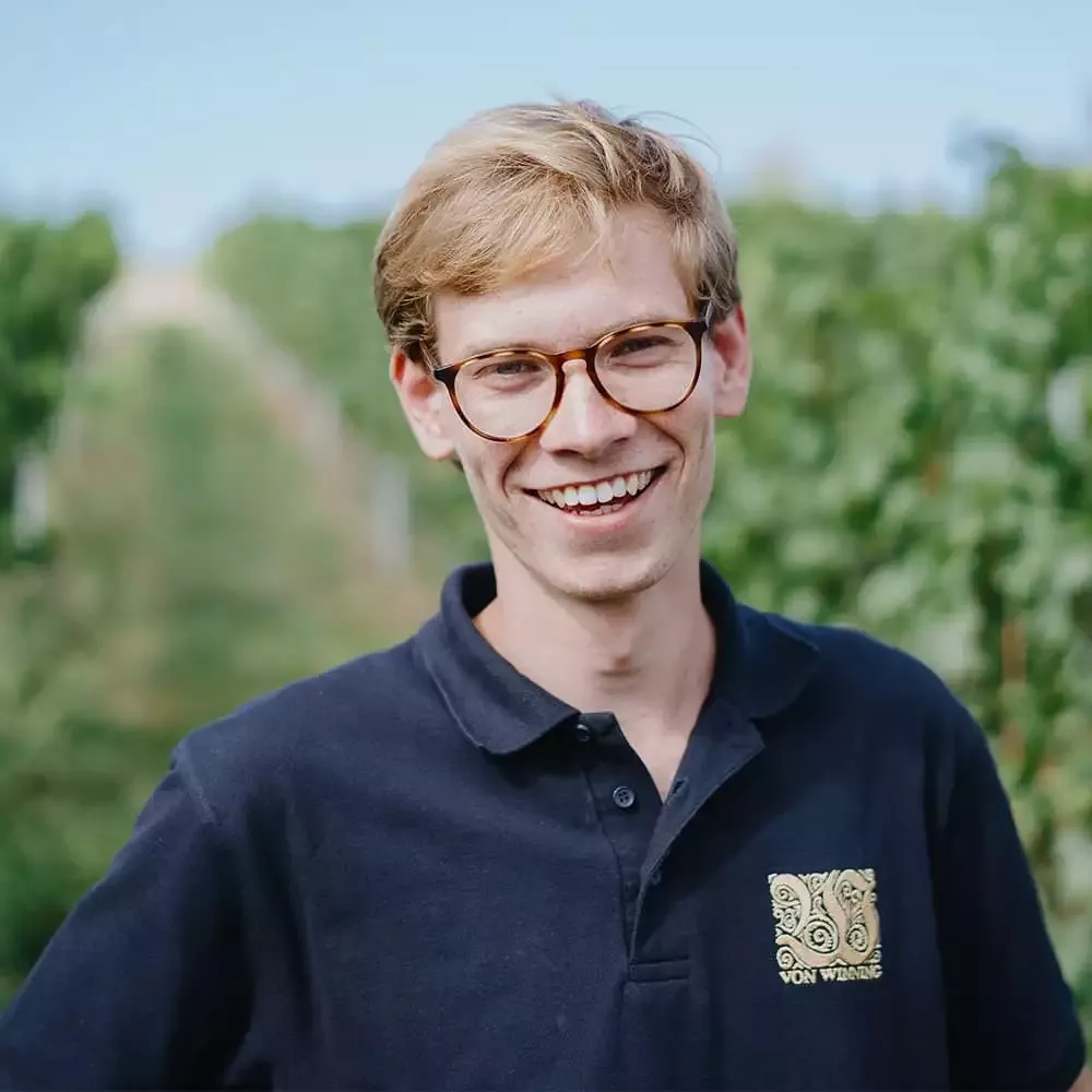 Eine lächelnde Person mit Brille und hellem Haar steht in einem Weinberg und trägt ein dunkles Poloshirt mit einem Logo. Im Hintergrund erstrecken sich die Weinreben unter einem klaren blauen Himmel.