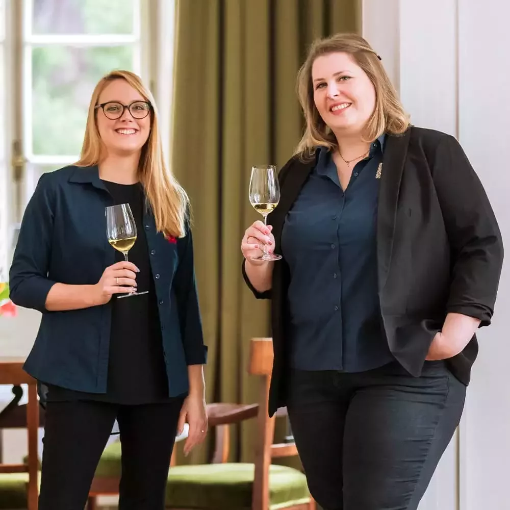 Zwei Frauen stehen lächelnd und mit Weingläsern in der Hand in einem hellen Raum. Beide tragen dunkle Hemden, eine trägt einen Blazer. Im Hintergrund sind ein Fenster und grüne Vorhänge zu sehen, außerdem ein Tisch und ein Stuhl. Sie wirken entspannt und lässig.