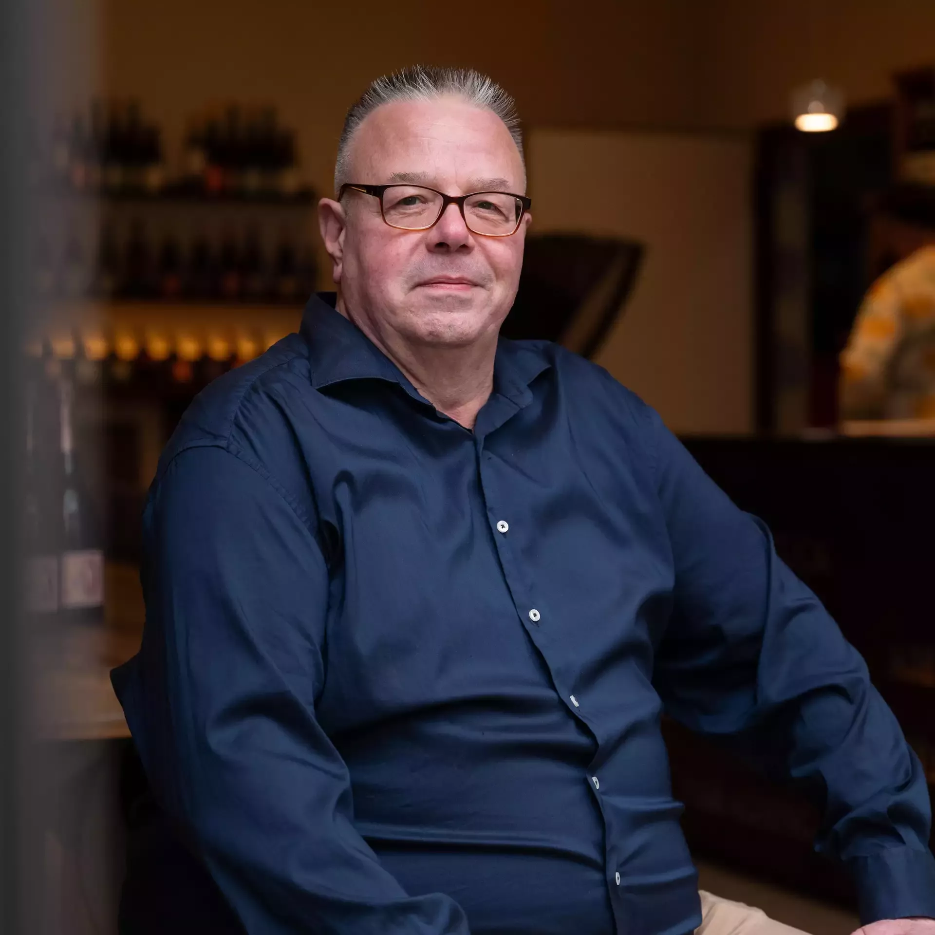 Ein Mann mit Brille und dunkelblauem Hemd sitzt in einem schwach beleuchteten Raum, im Hintergrund stehen Flaschen auf Regalen und blickt in die Kamera.