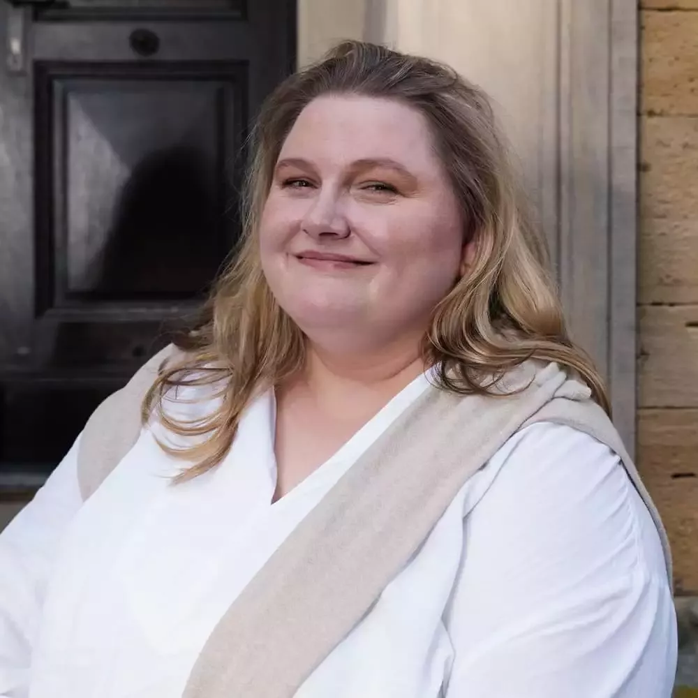 Eine Person mit langen blonden Haaren, die ein weißes Hemd und eine beige Strickjacke trägt, lächelt sanft, während sie draußen vor einer Holztür steht. Im Hintergrund ist eine Steinmauer zu sehen.