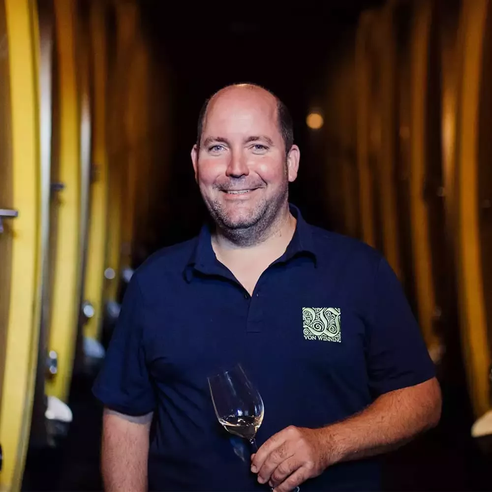 Ein lächelnder Mann hält ein Weinglas in einem schwach beleuchteten Weingut mit großen Holzfässern im Hintergrund. Er trägt ein marineblaues Hemd mit einem Logo auf der Brust.