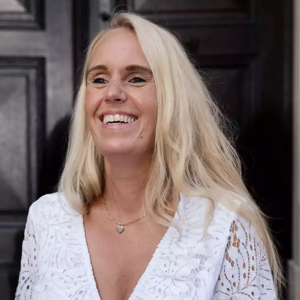 Eine Person mit langen blonden Haaren lächelt warmherzig. Sie trägt eine weiße, strukturierte Bluse und eine Halskette mit herzförmigem Anhänger. Im Hintergrund ist eine dunkle Holztür zu sehen.
