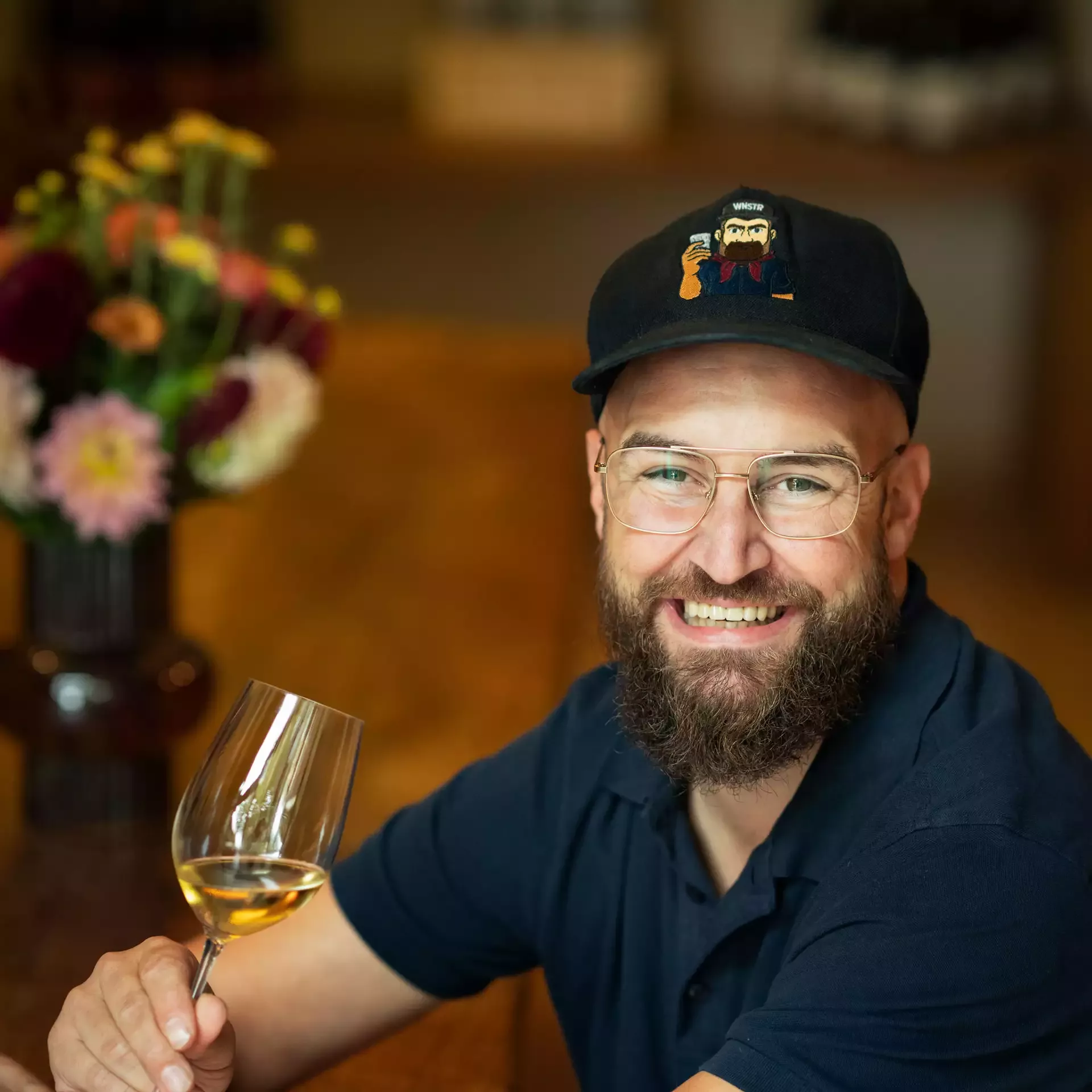 Ein bärtiger Mann mit schwarzer Mütze und Brille lächelt, während er ein Glas Weißwein hält. Er sitzt an einem Holztisch mit einem verschwommenen Blumenarrangement im Hintergrund.
