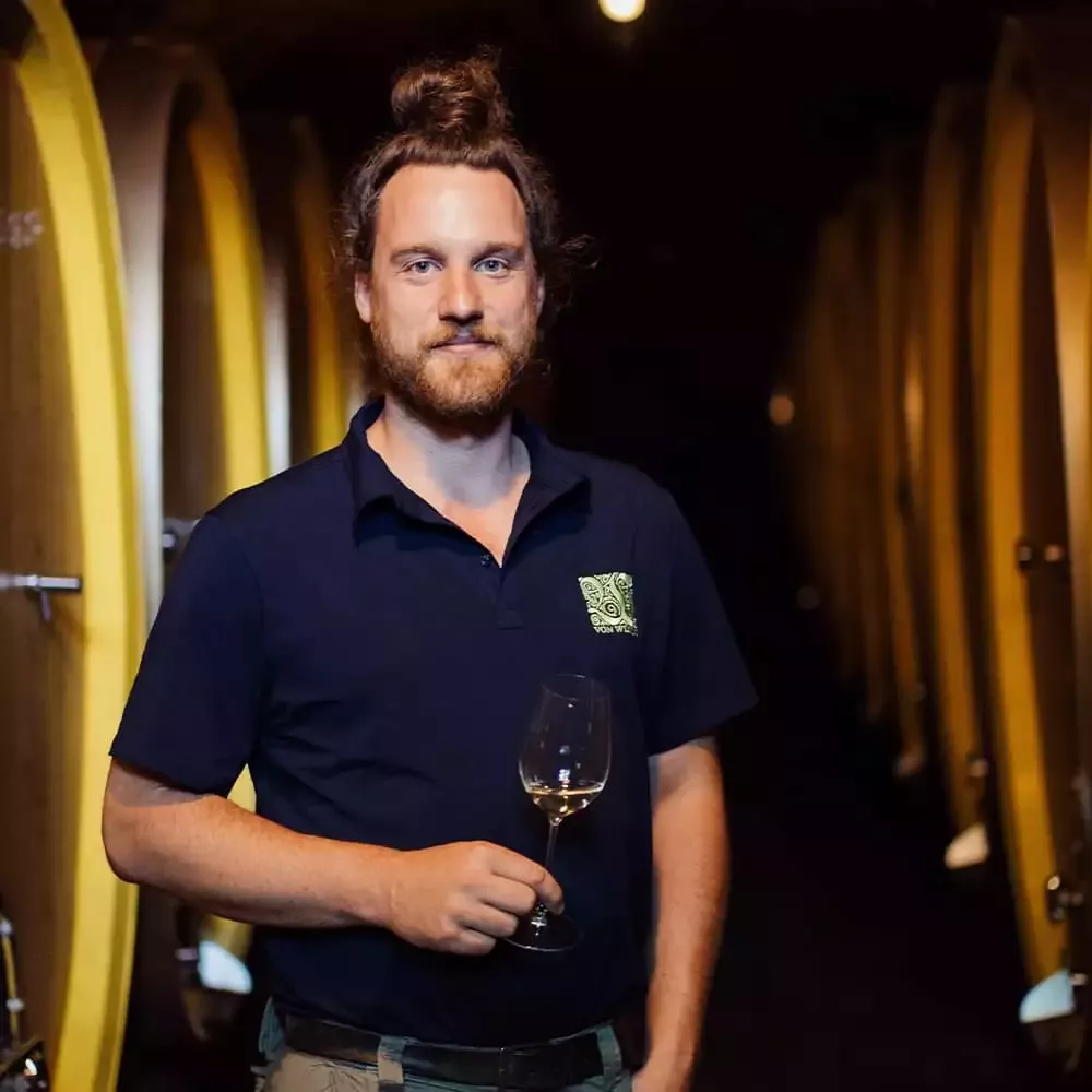 Mann mit Bart und Haarknoten in der Hand steht in einem schwach beleuchteten Keller, der von großen Holzfässern umgeben ist. Er trägt ein marineblaues Poloshirt mit einem Logo darauf.