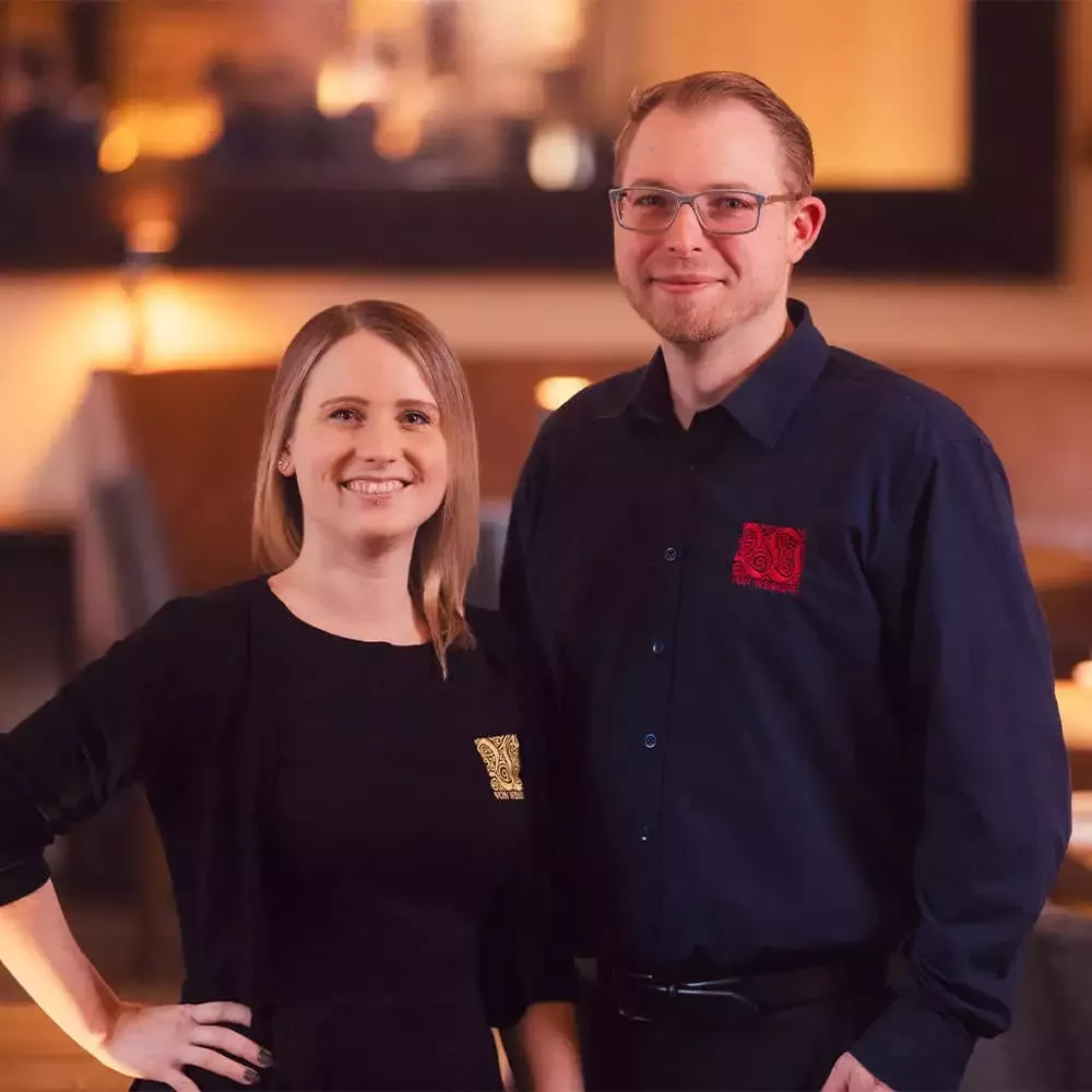 Ein Mann und eine Frau stehen nebeneinander und lächeln. Der Mann trägt eine Brille und ein dunkles Hemd mit rotem Logo, während die Frau ein schwarzes Oberteil mit einem hellen Logo trägt. Der Hintergrund ist sanft beleuchtet.