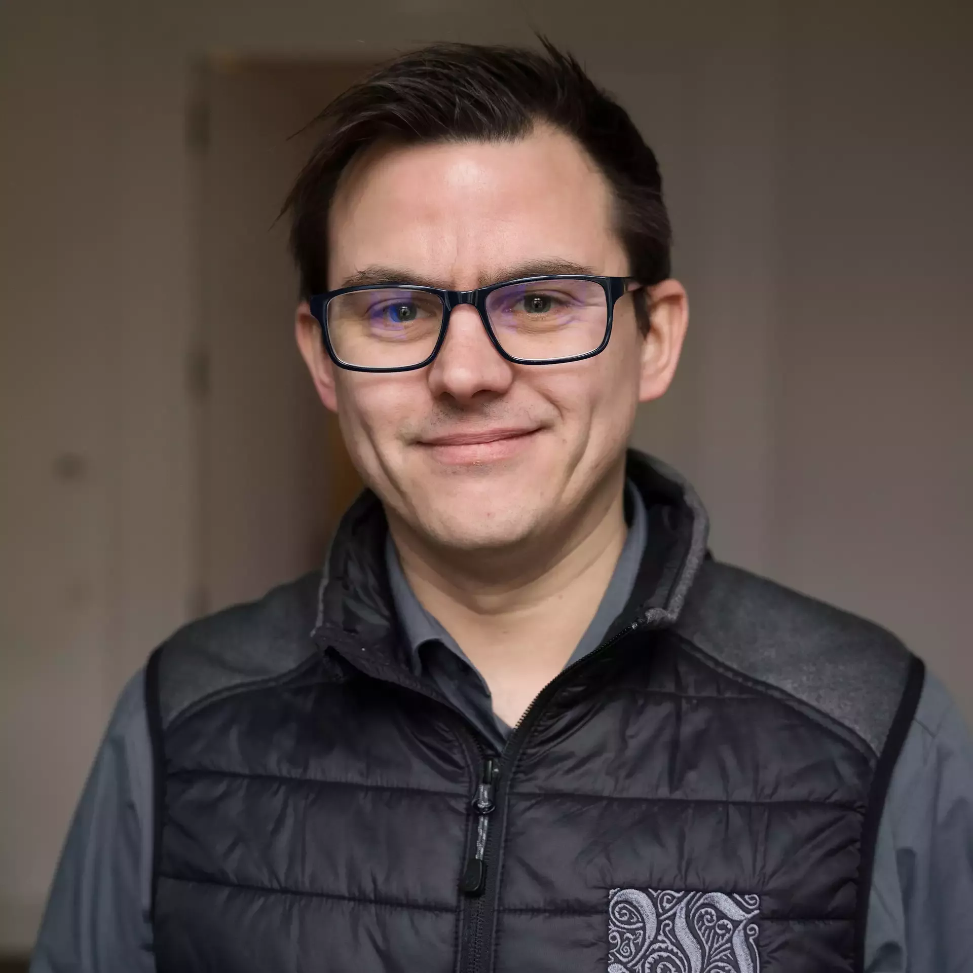 Eine Person mit kurzen Haaren und Brille lächelt in die Kamera. Sie trägt eine schwarze Steppweste über einem Hemd mit Knöpfen. Der Hintergrund ist ein sanft beleuchteter, neutral gefärbter Innenraum.