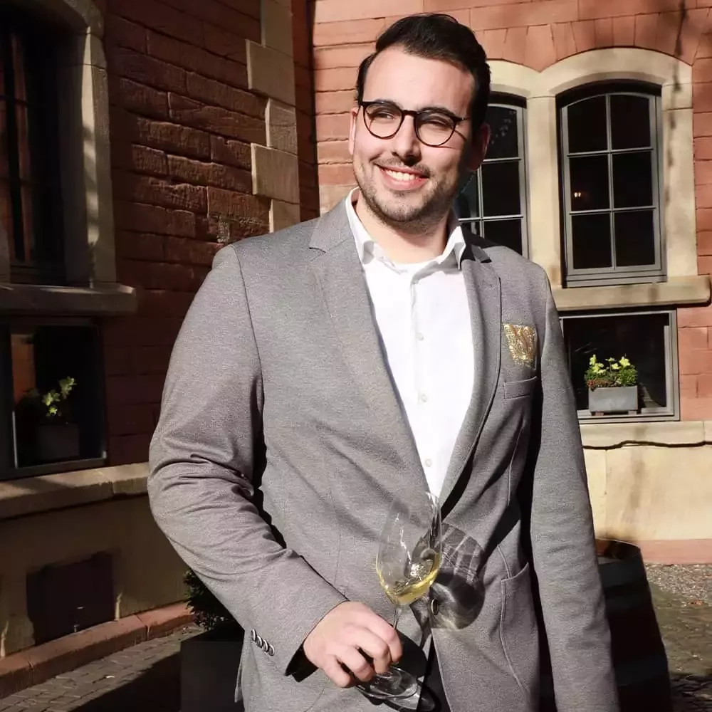 Ein Mann mit dunklem Haar und Brille steht draußen im Sonnenlicht. Er trägt einen hellgrauen Blazer und ein weißes Hemd. Er hält ein Weinglas und lächelt. Im Hintergrund sind ein Backsteingebäude und große Fenster zu sehen.