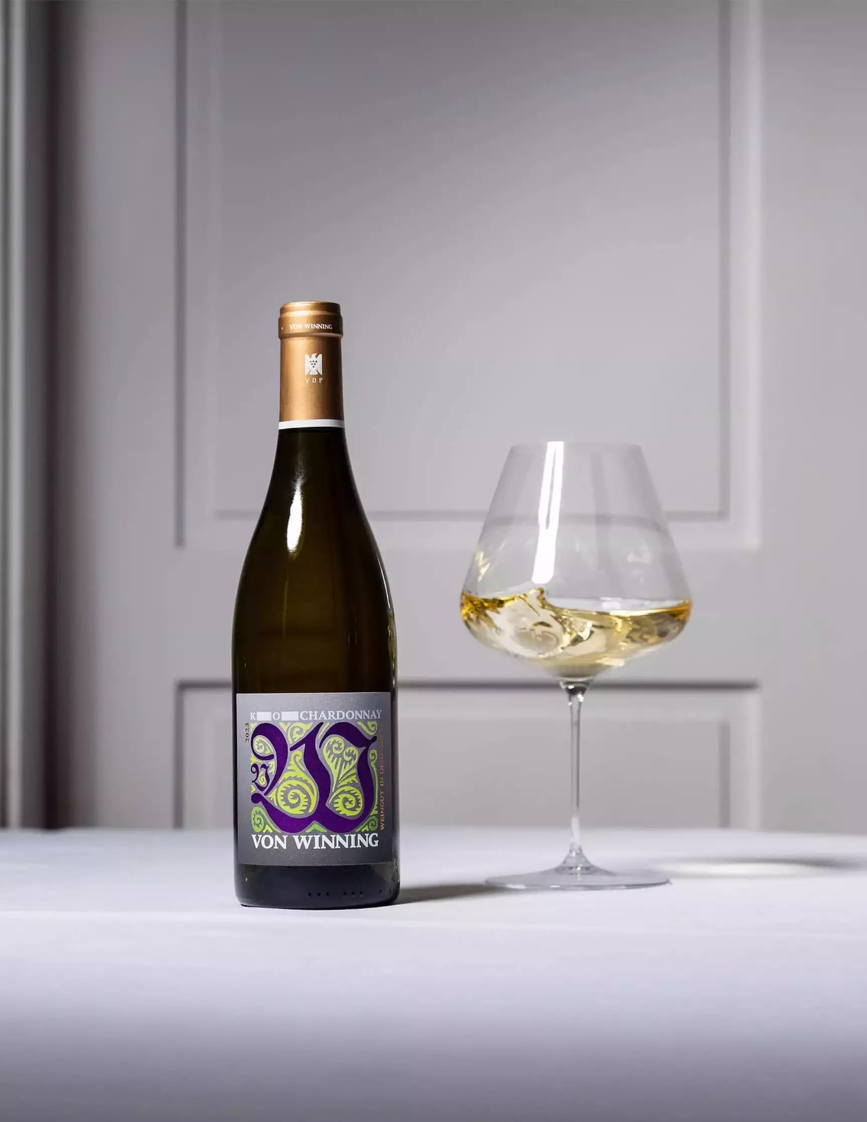 Eine Flasche Von Winning 2020 Chardonnay steht neben einem mit Weißwein gefüllten Weinglas auf einer weißen Tischdecke. Die Flasche zeigt ein farbenfrohes, abstraktes Etikettendesign. Der Hintergrund ist eine schlichte, hellgraue Wand mit dezenter Verkleidung.