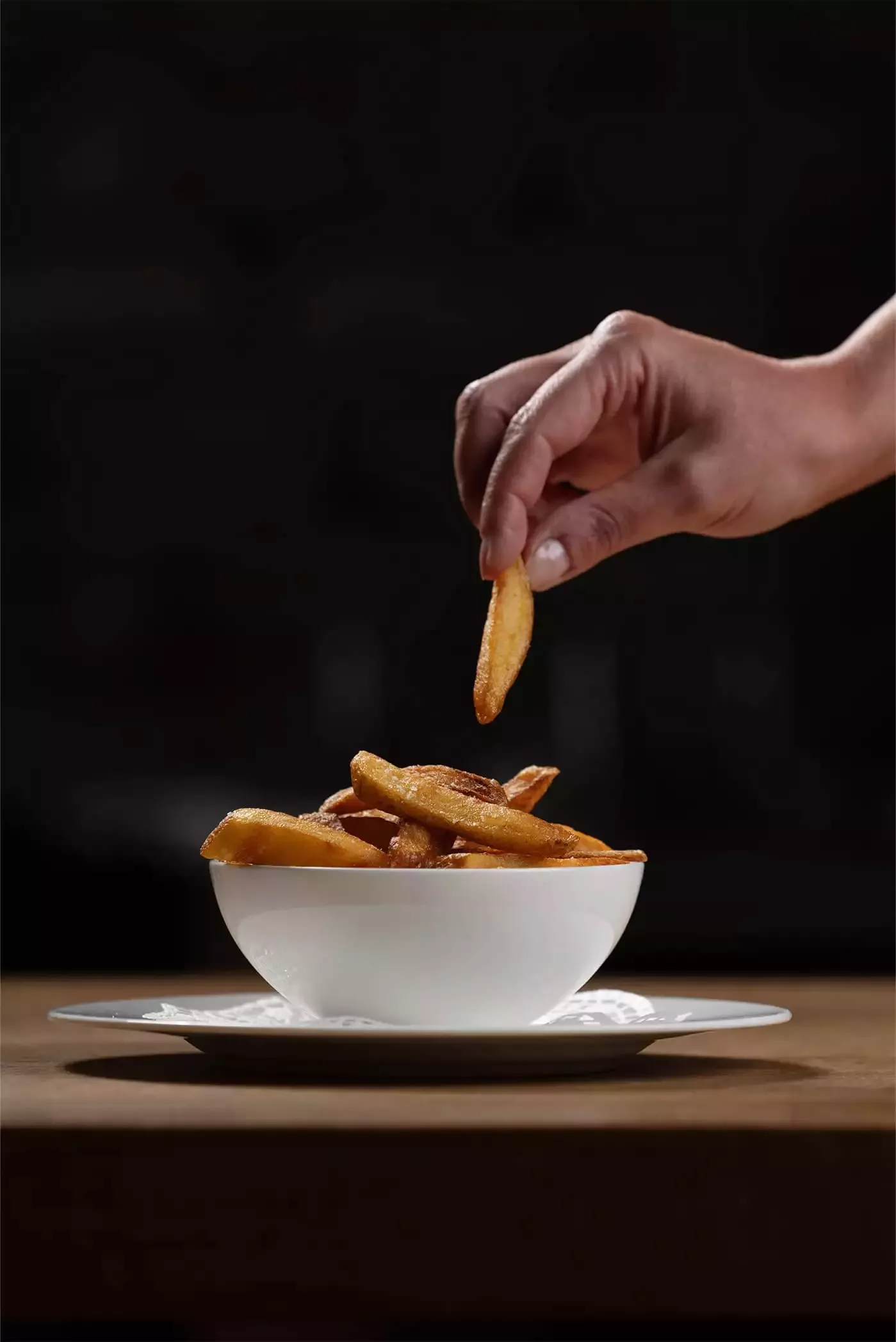Eine Hand nimmt eine Pommes Frites aus einer Schüssel mit goldbraunen, knusprigen Pommes Frites. Die Schüssel steht auf einem Teller vor einem dunklen Hintergrund.