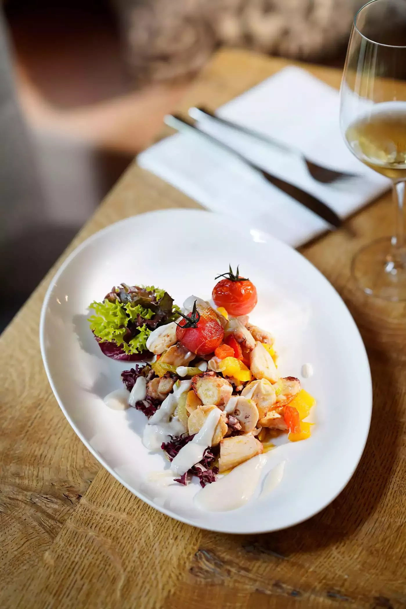 Ein weißer Teller mit Meeresfrüchtesalat, garniert mit zwei Kirschtomaten, Salat an der Seite, beträufelt mit weißem Dressing. Im Hintergrund auf einem Holztisch stehen ein Glas Weißwein und Besteck.