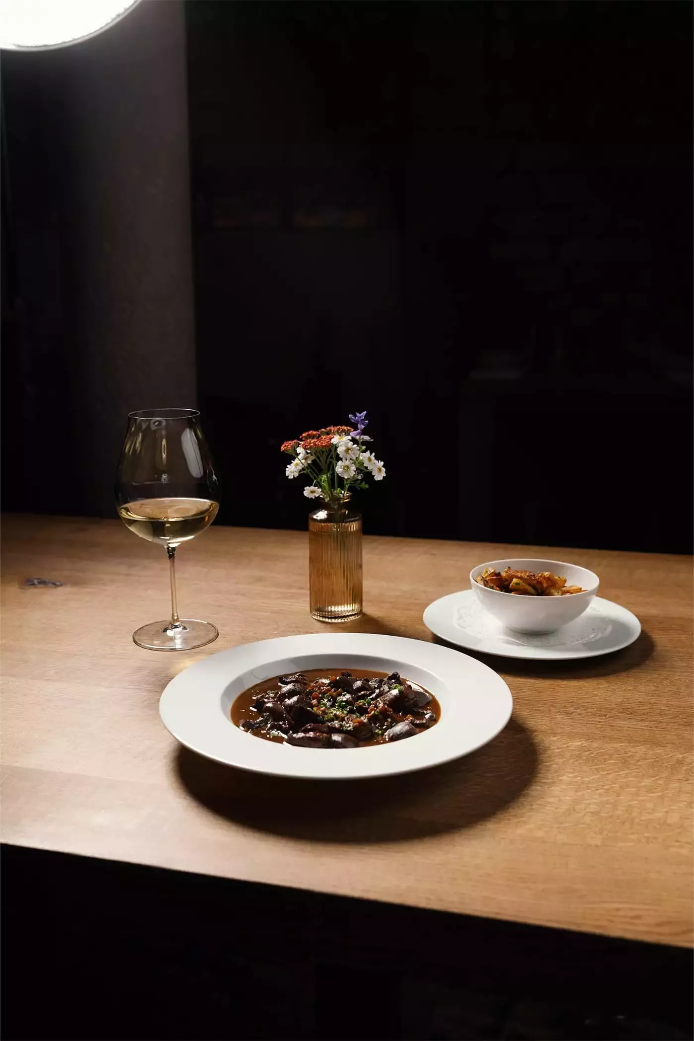 [Translate to English:] Holztisch mit stilvoll angerichteten Essen auf einem weißen Teller, Schüssel Bratkartoffeln, Weinglas und dekorativen Blumen