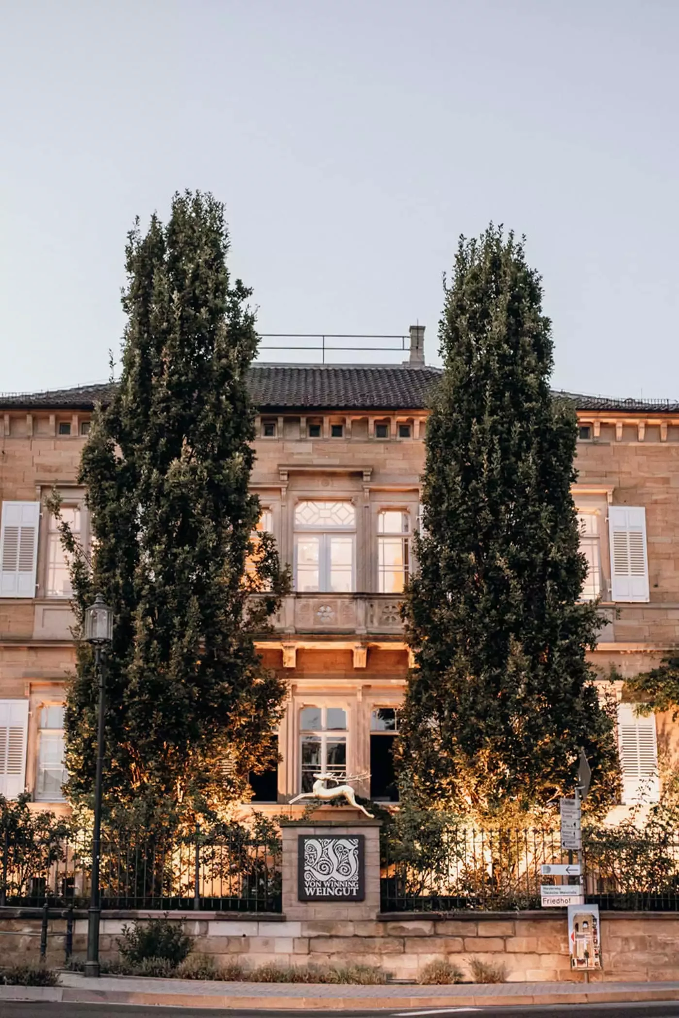 Ein stattliches, zweistöckiges Steingebäude mit hohen Fenstern und Fensterläden, flankiert von zwei hohen Bäumen. Die Fassade ist warm beleuchtet, wodurch die architektonischen Details des Gebäudes hervorgehoben werden. Vor dem Anwesen ist ein dekoratives Schild mit der Aufschrift Weingut Von Winning zu sehen.