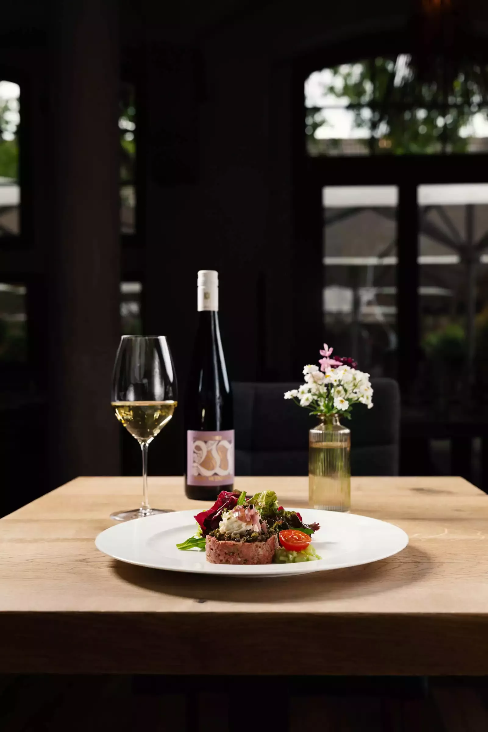 Ein Holztisch mit einem Teller Gourmet-Essen, darunter ein Fleischgericht, Gemüse und Kirschtomaten. Daneben stehen ein Weinglas und eine Flasche Weißwein. Im Hintergrund steht eine kleine Blumenvase, dahinter sind verschwommene Fenster.