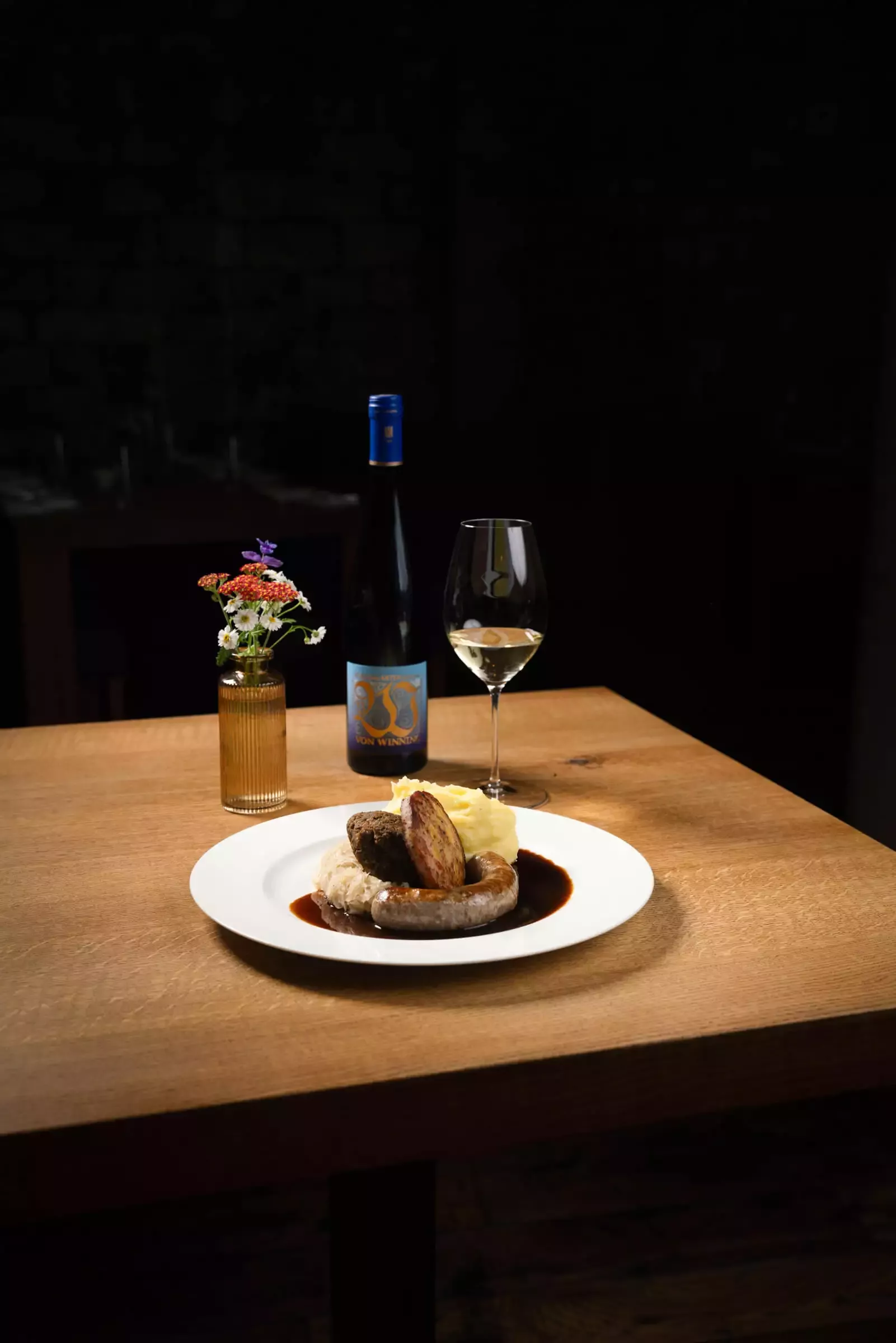 Auf einem Holztisch steht ein Teller mit Gourmetwürsten in Soße. Daneben stehen eine Flasche Wein und ein Glas Weißwein. Auf dem Tisch steht vor einem dunklen Hintergrund auch eine kleine Vase mit bunten Blumen.
