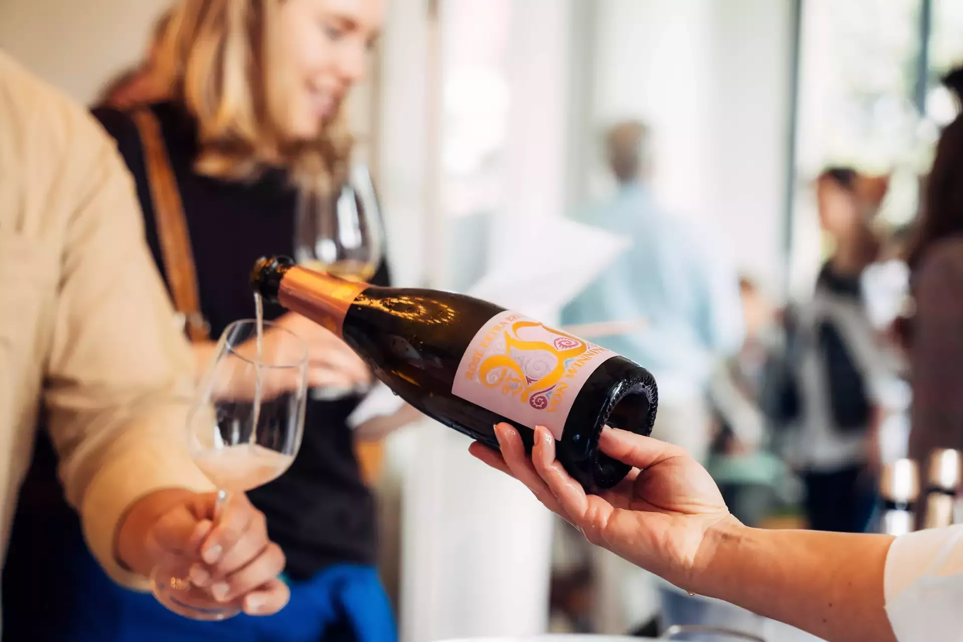 Von Winning Rosé-Sekt wird bei einer Verkostung in ein Glas eingeschenkt, im Hintergrund Personen in Gespräch.