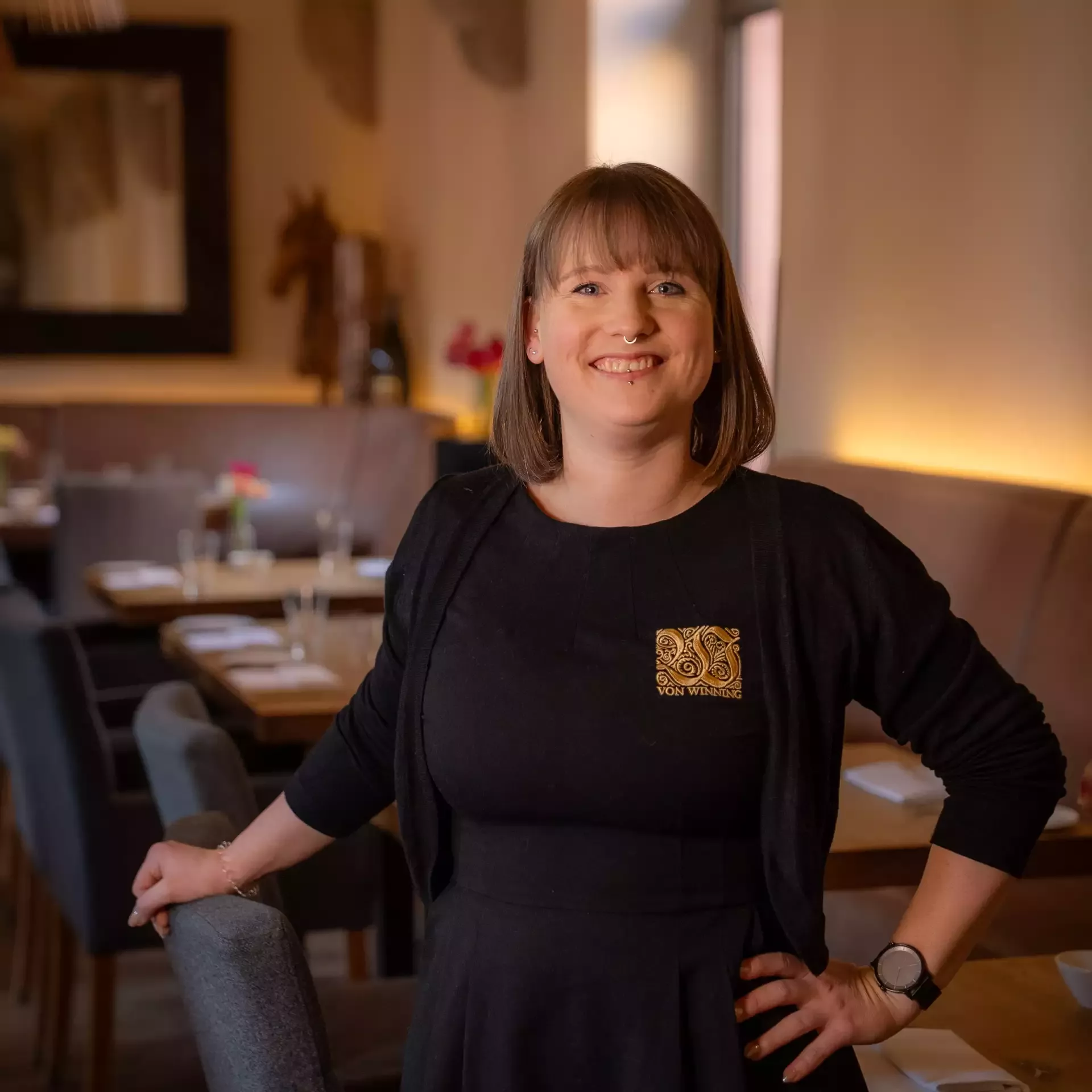 Lächelnde Frau im schwarzen Kleid steht in einem warm beleuchteten Restaurant neben gedeckten Tischen.