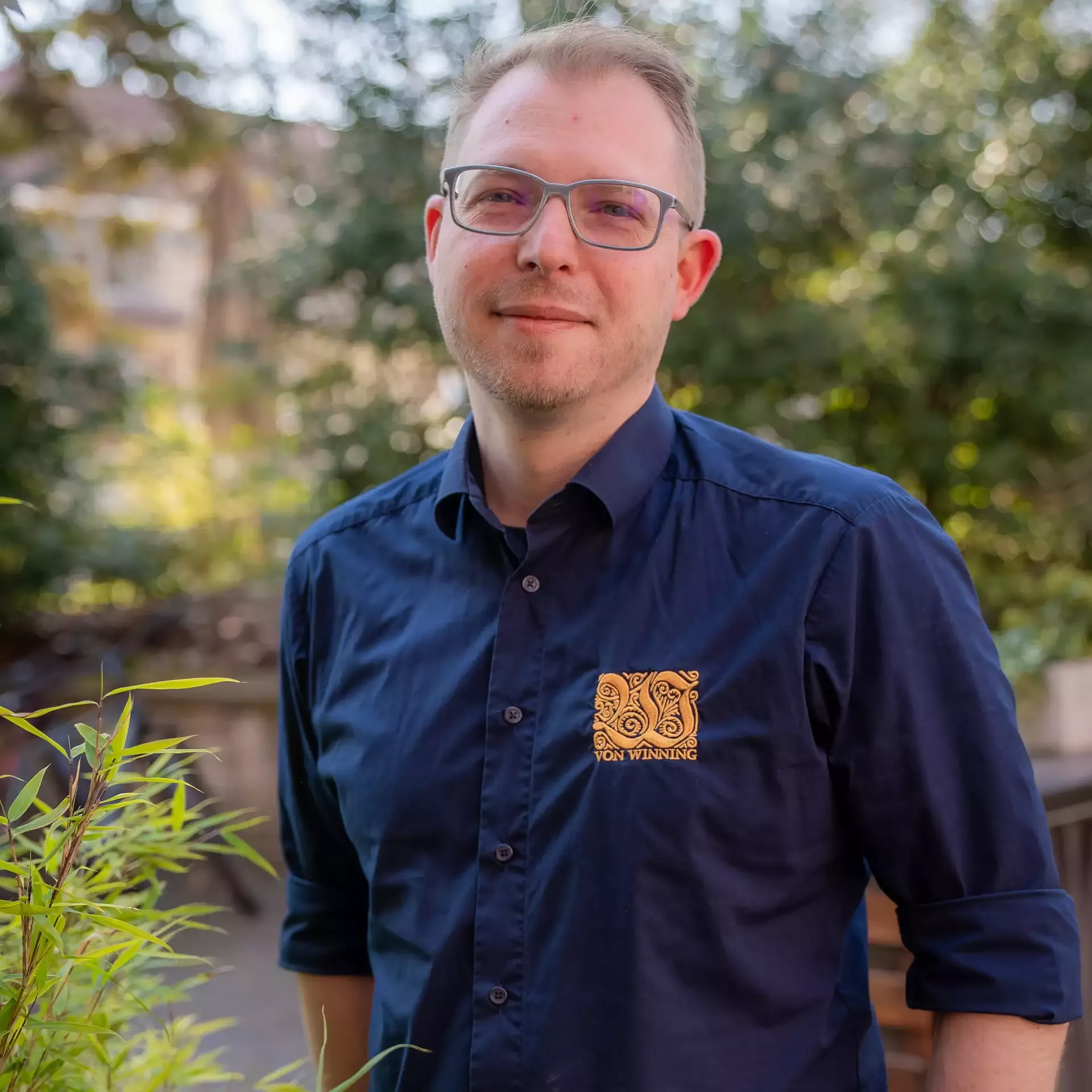 Mann mit Brille und dunkelblauem Hemd steht entspannt im Freien vor unscharfem grünen Hintergrund.