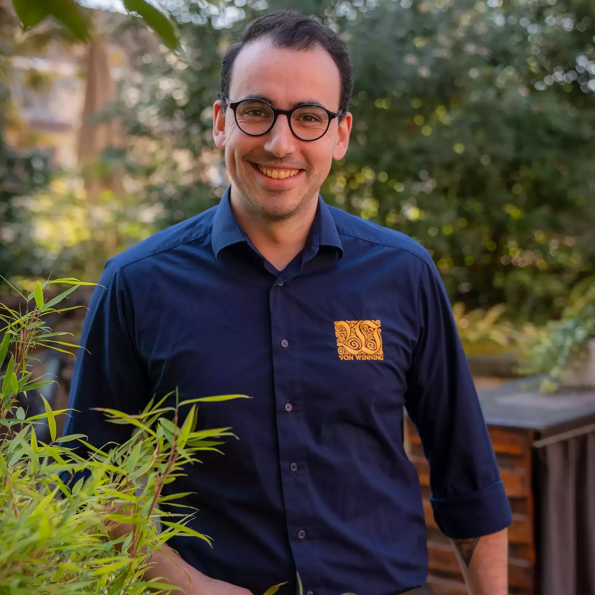Lächelnder Mann mit Brille und dunkelblauem Hemd steht draußen zwischen grünen Pflanzen in einem Garten.