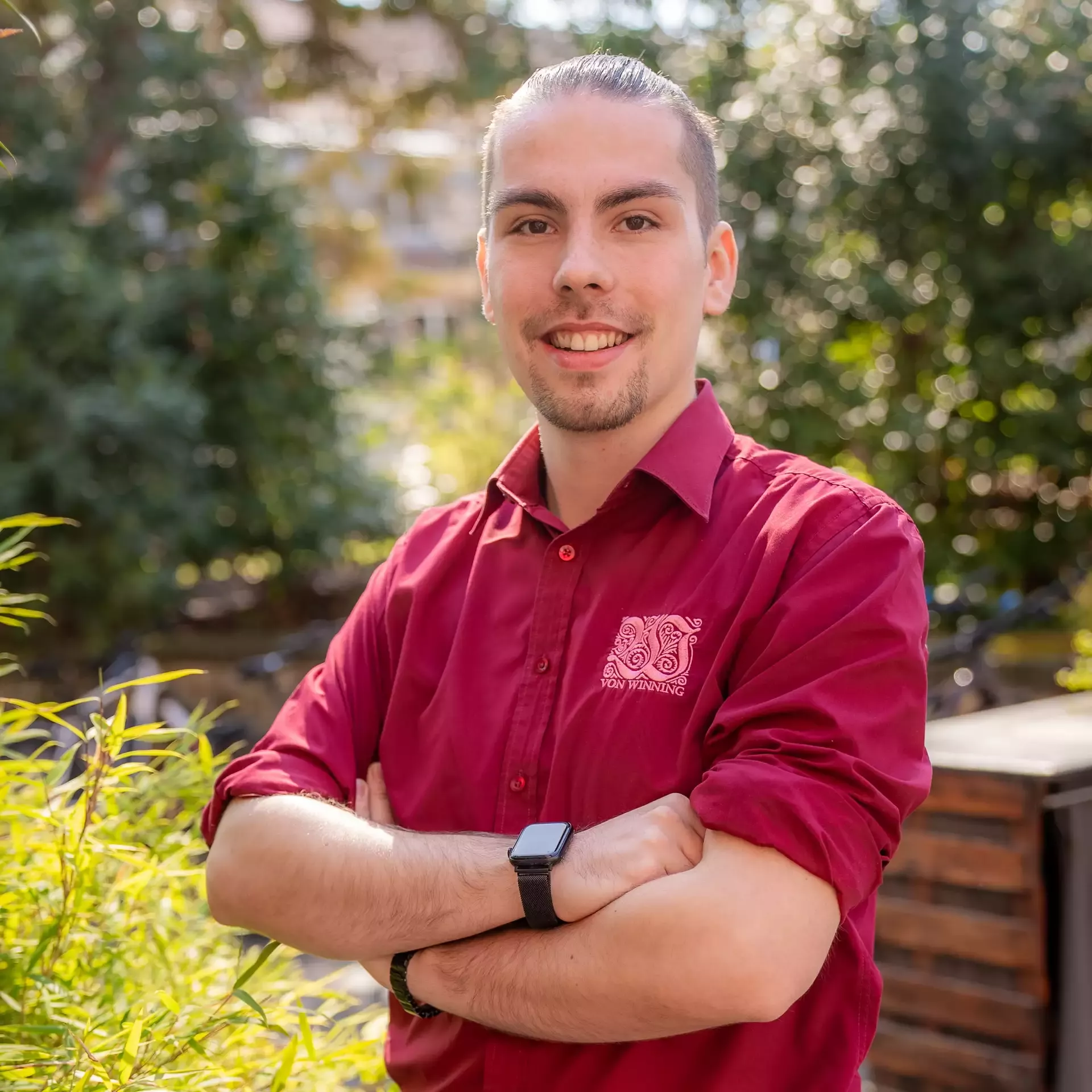 Mitarbeiter des Weinguts Von Winning in rotem Hemd steht mit verschränkten Armen im Garten und lächelt in die Kamera.